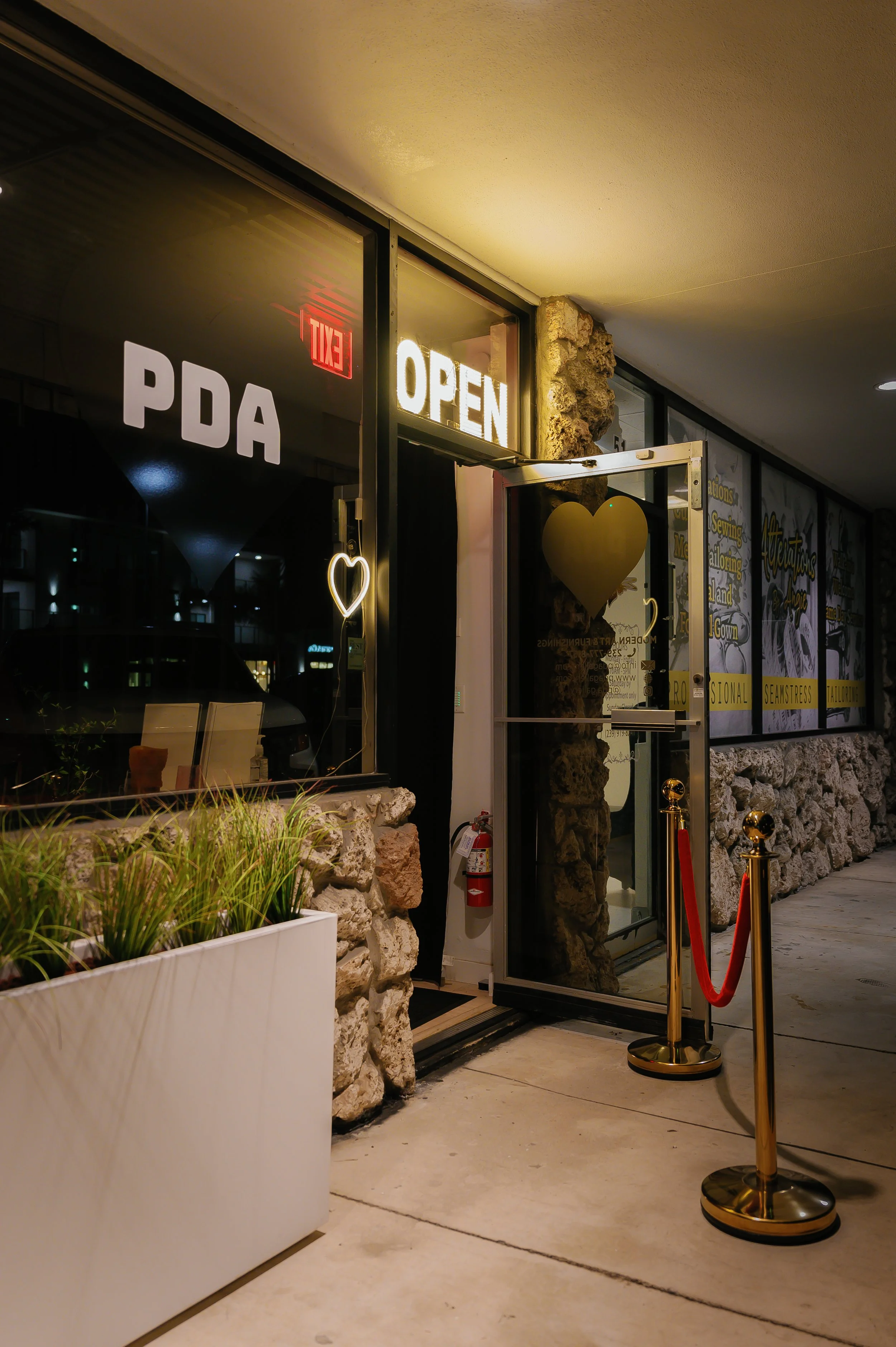 Nighttime exterior of a storefront with illuminated 'OPEN' sign and white 'PDA' lettering on window. Roped-off entrance with gold stanchions, a fire extinguisher nearby, and stone wall accents. Potted plants in the foreground.