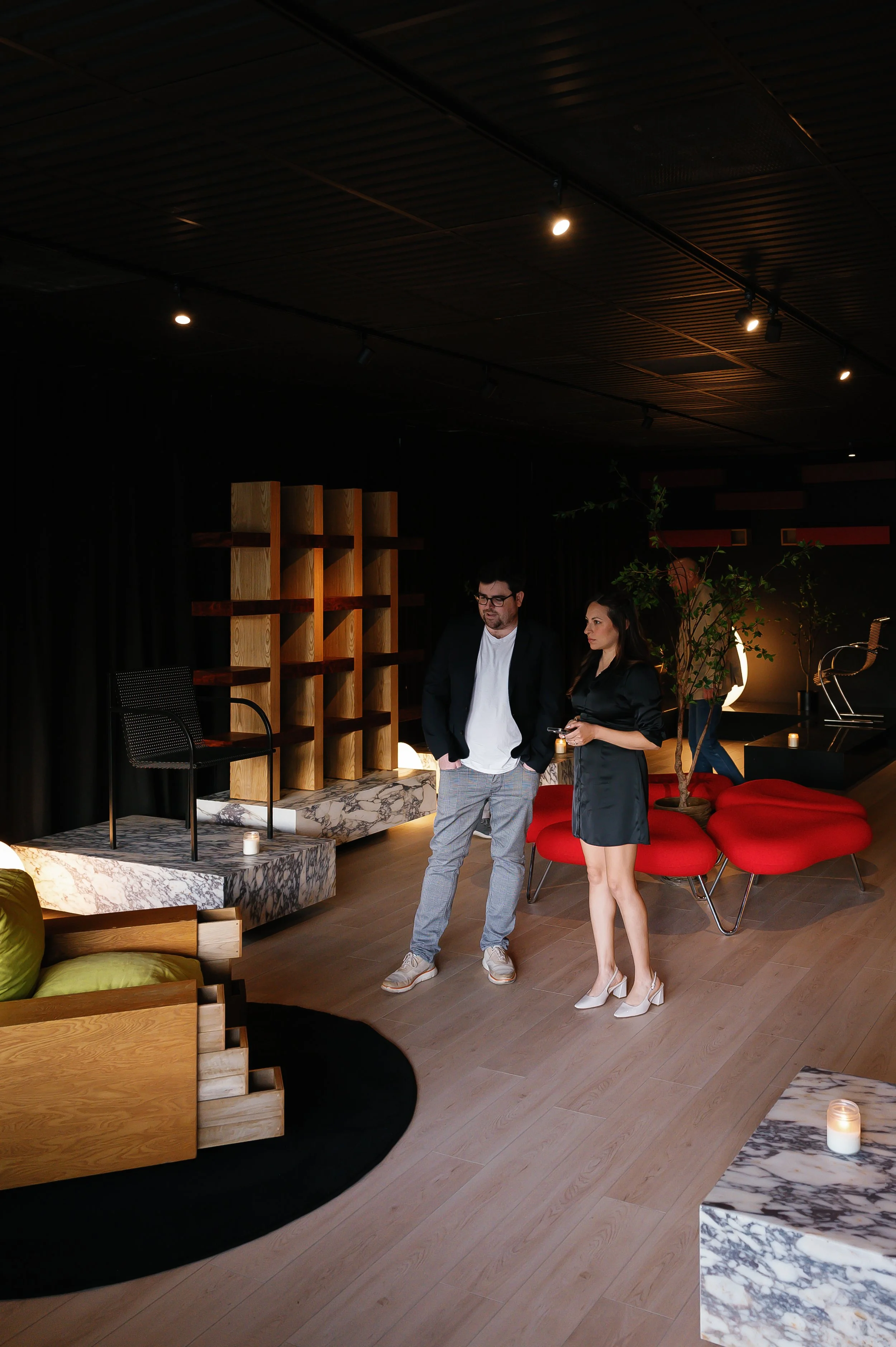 Two people standing in a modern, dimly lit lounge area with contemporary furniture, candles, and potted plants.
