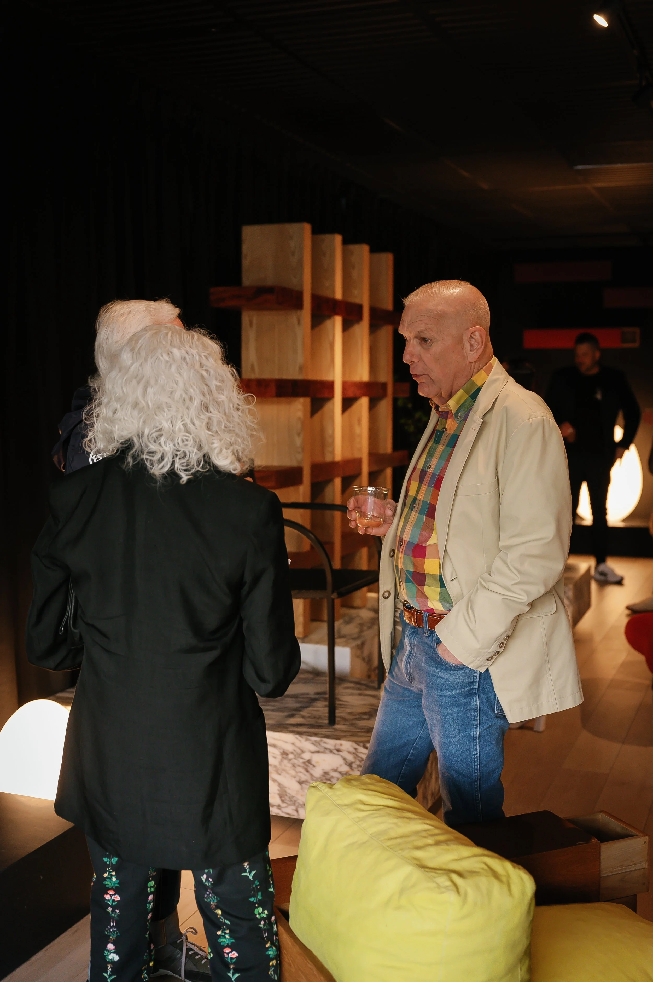 Two people having a conversation in a dimly lit room, one holding a glass of drink.