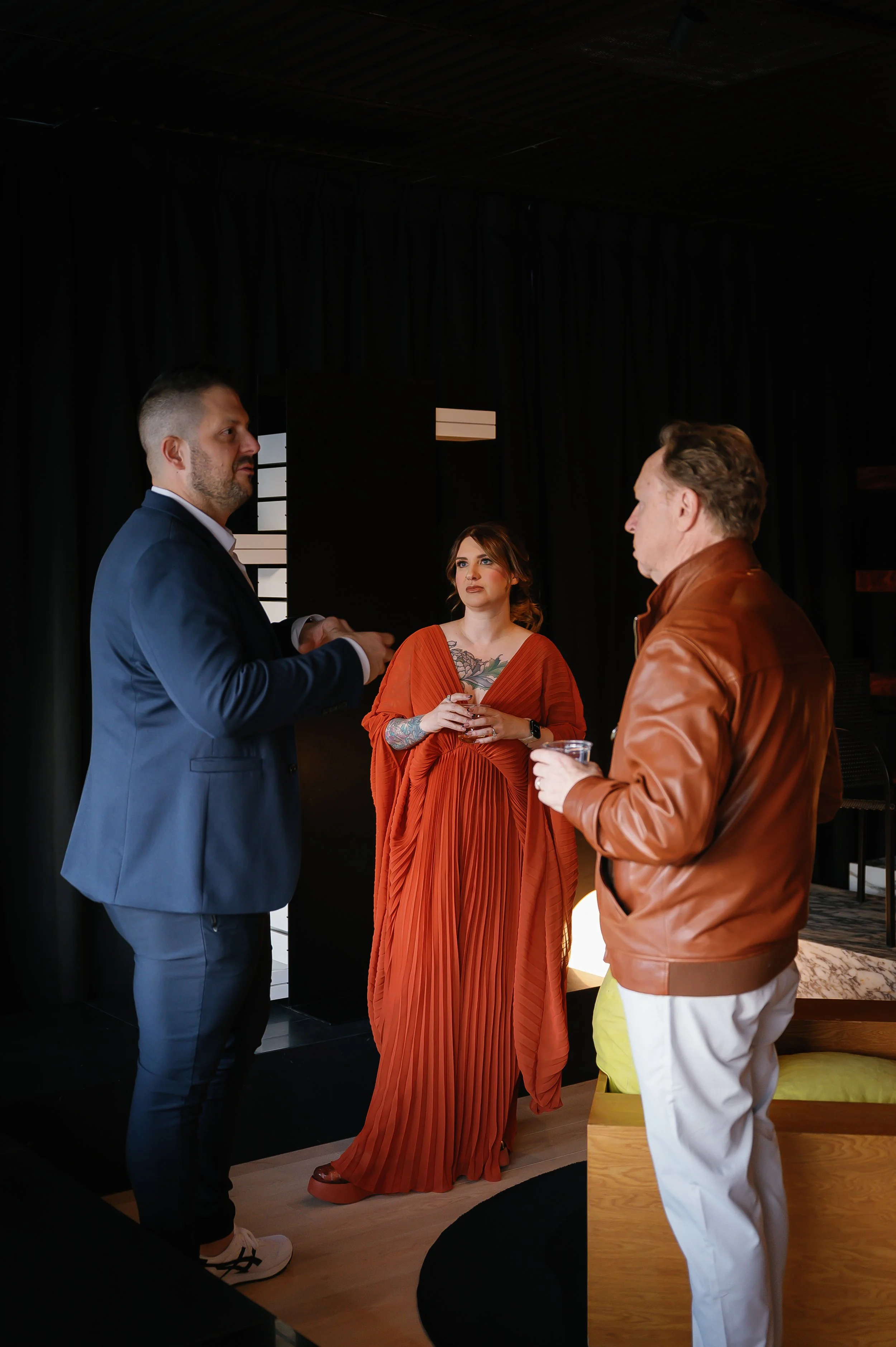 Three people, two men and one woman, are engaged in a conversation indoors with dark walls and minimal decor. The man on the left wears a navy suit and white sneakers, the woman in the center wears an orange pleated gown with tattoos visible on her c