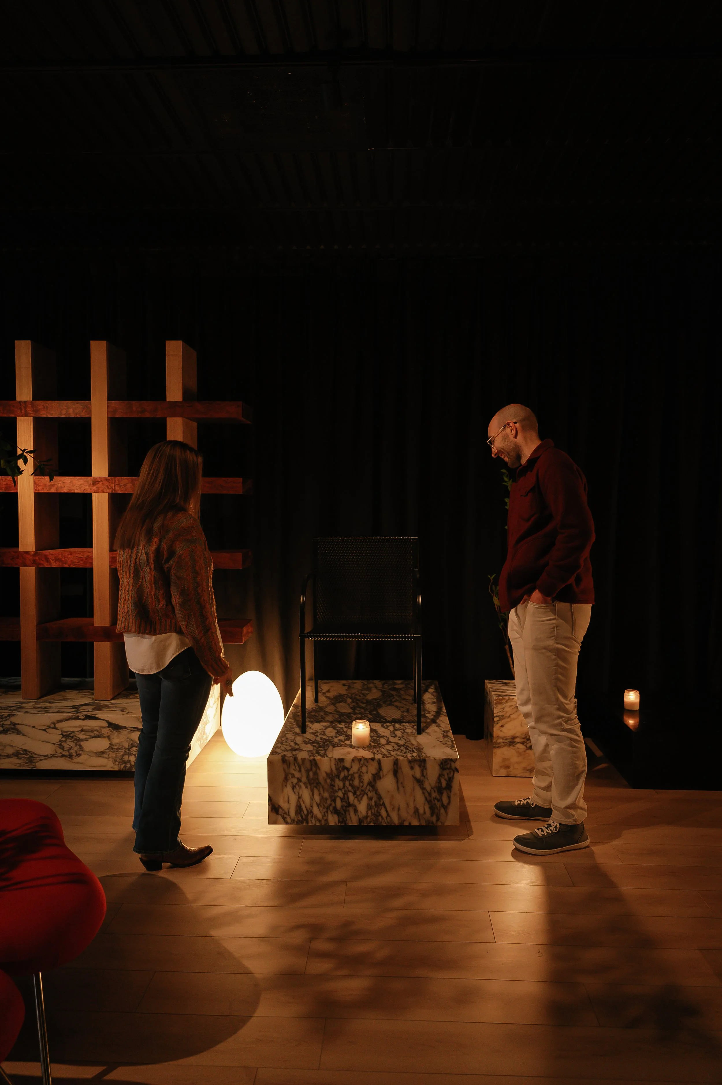 A woman and man standing in a dimly lit room with a black curtain, marble pedestal, candles, and a black chair, engaging in conversation.