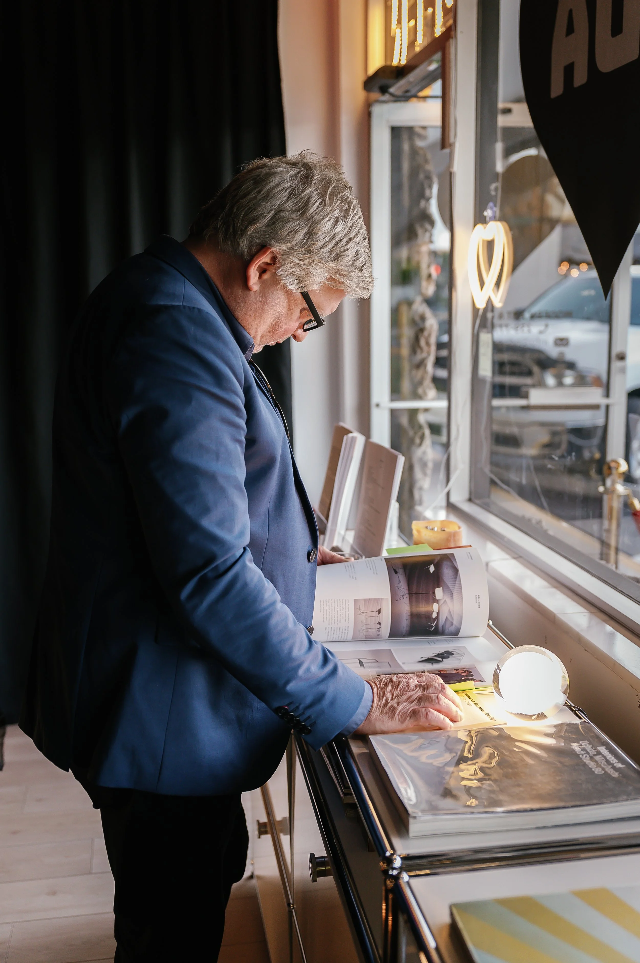 A man with gray hair and glasses, wearing a blue blazer, browsing magazines and books displayed on a table by a window in a shop.