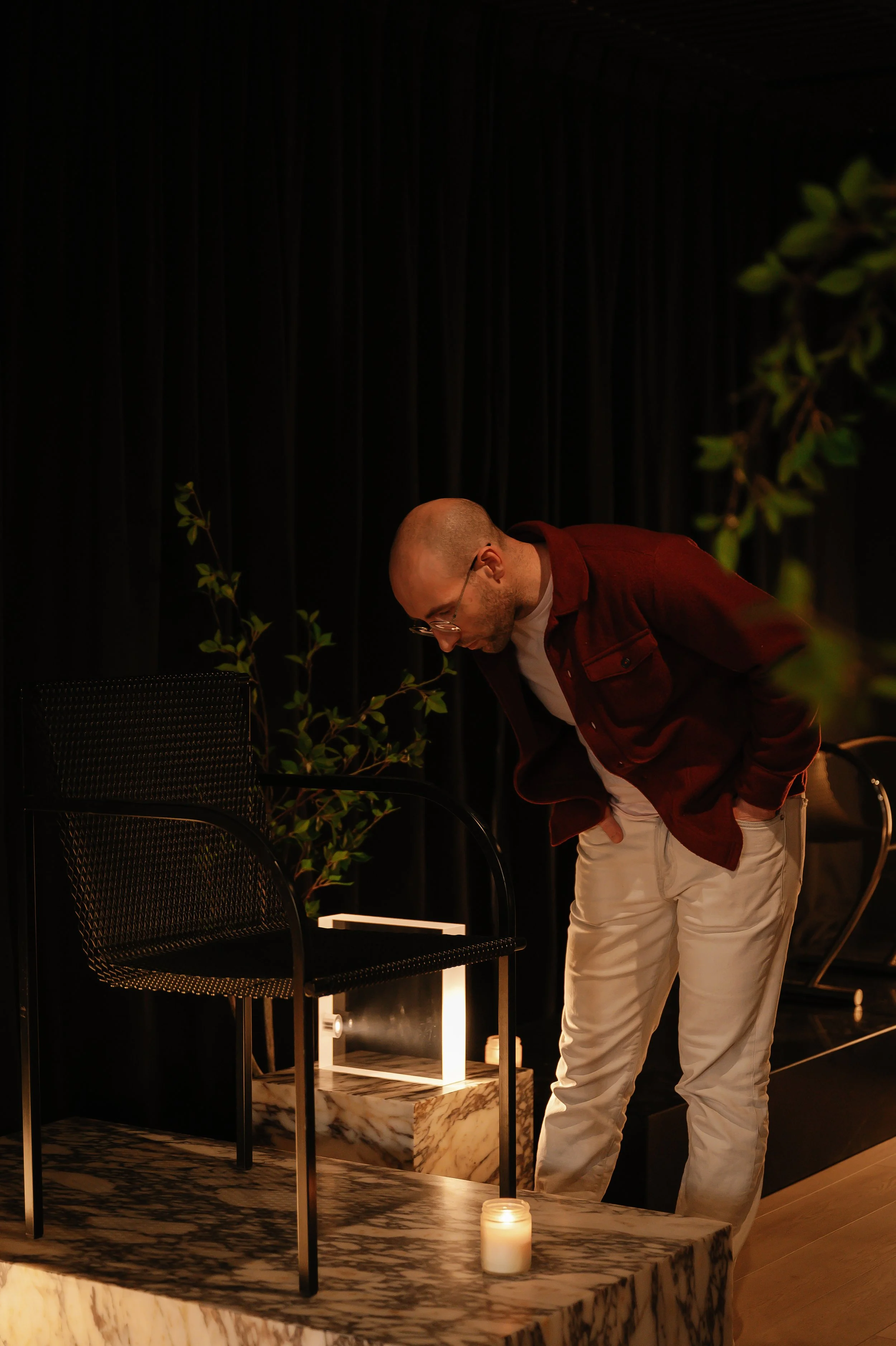 A man with glasses and a bald head bowing his head in an indoor setting. There are plants, a candle, and a black chair on a marble surface.