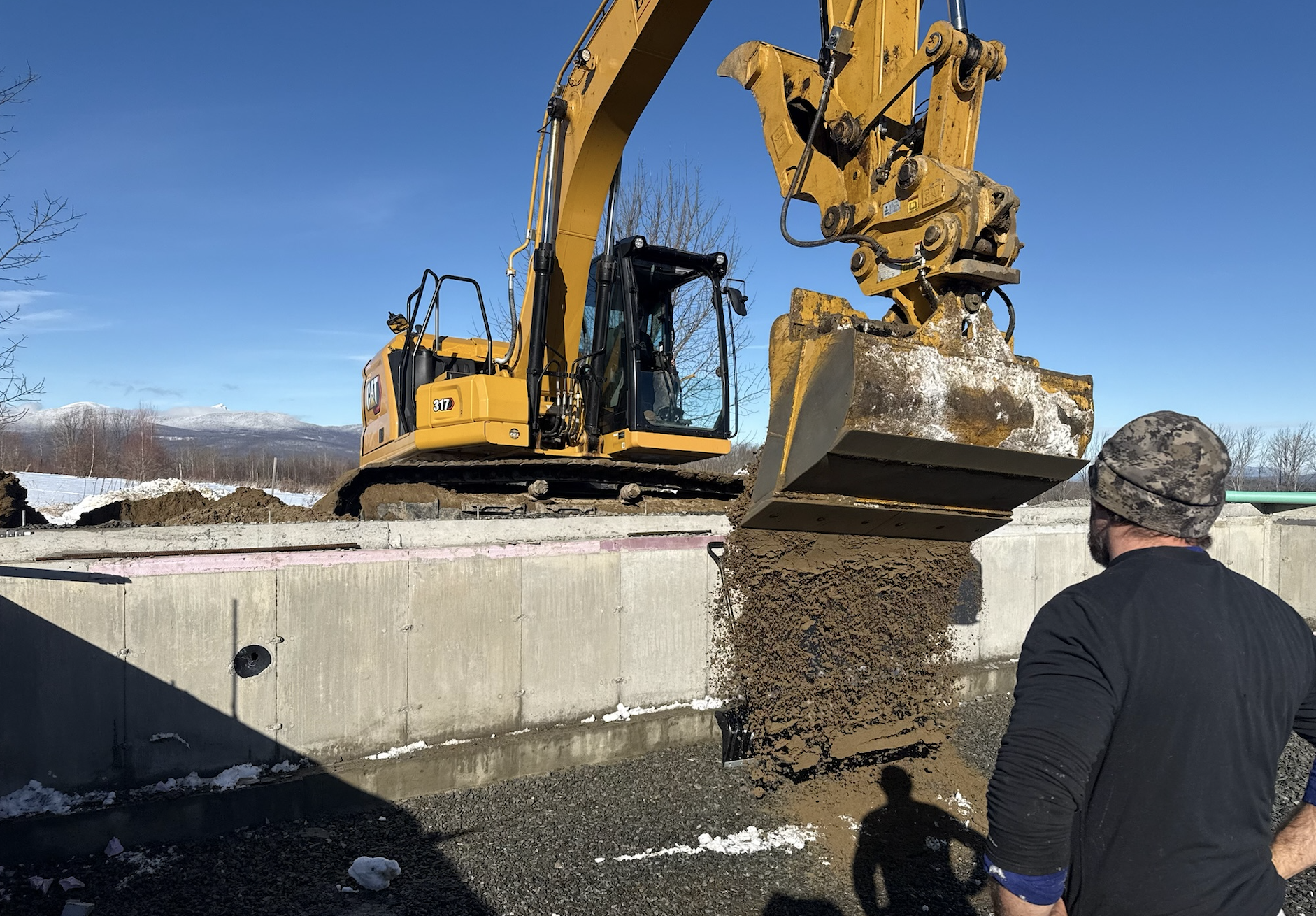 A yellow excavator pouring dirt into a trench with a man observing nearby, snow on the ground, and mountains in the background under a clear blue sky.