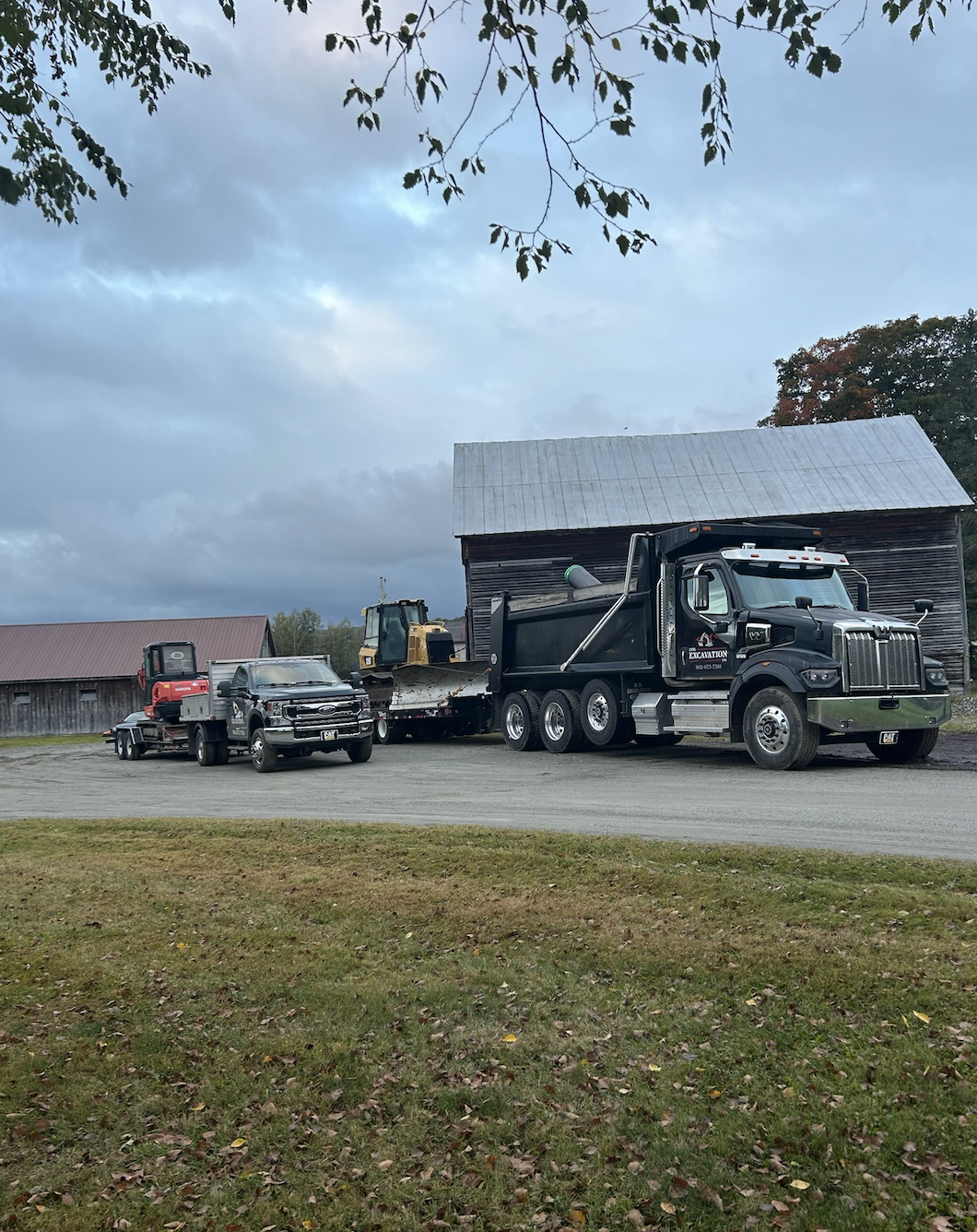 A construction site with a large black dump truck, a yellow excavator, a smaller red and black truck, and a silver pickup truck. There is a rustic barn in the background under a cloudy sky, and the scene is framed by overhanging tree branches.