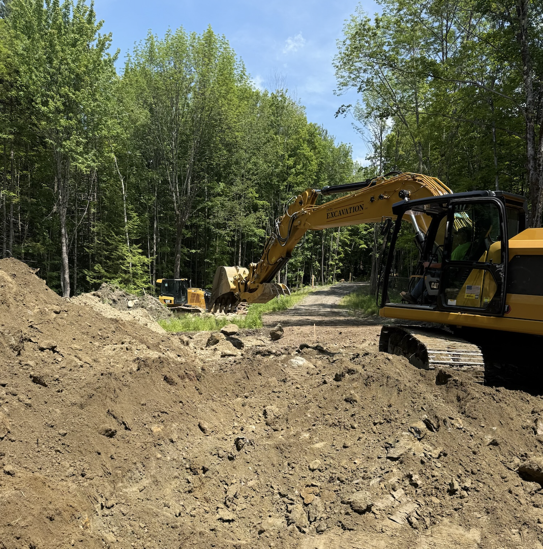 Construction site with a yellow excavator digging dirt along a forested dirt road, with trees and blue sky in the background.