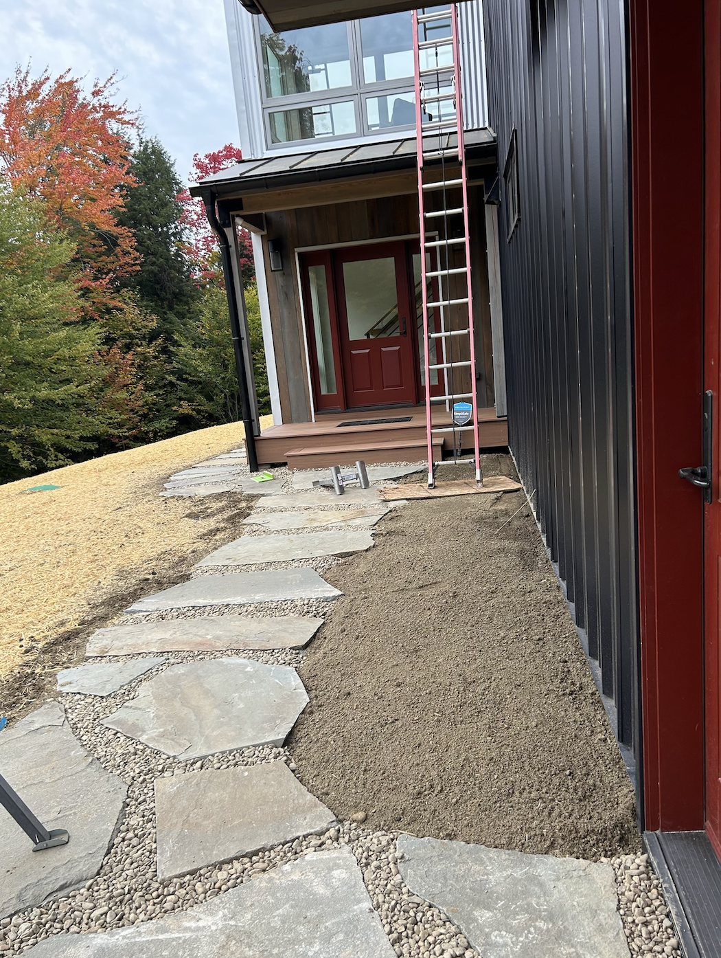 Stone pathway leading to a front porch with a red door, under construction with a ladder and construction materials nearby. Trees with autumn foliage in the background.
