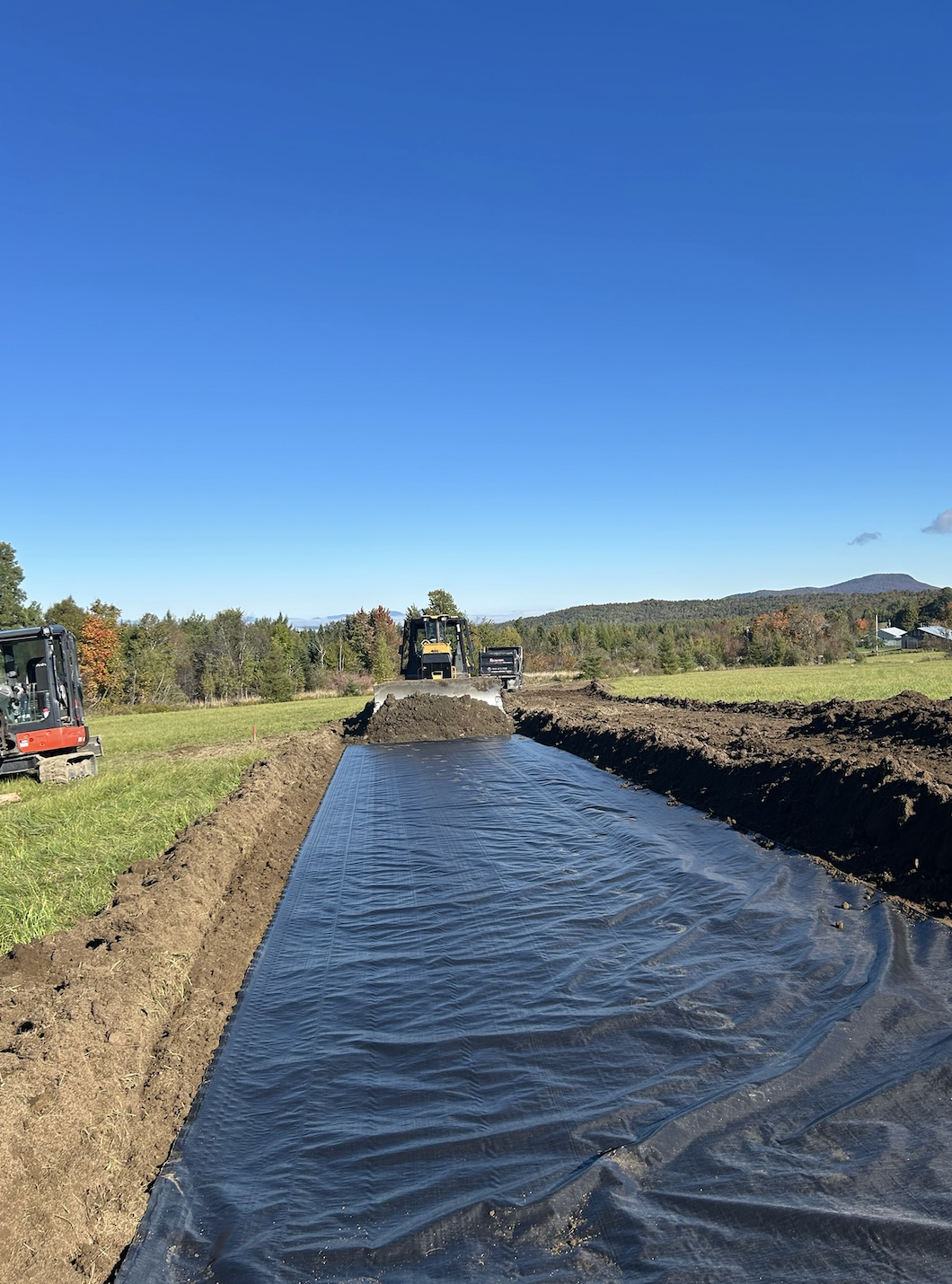 Construction site with a large black plastic sheet laid on the ground, excavators working in a field with trees and mountains in the distance under a clear blue sky.