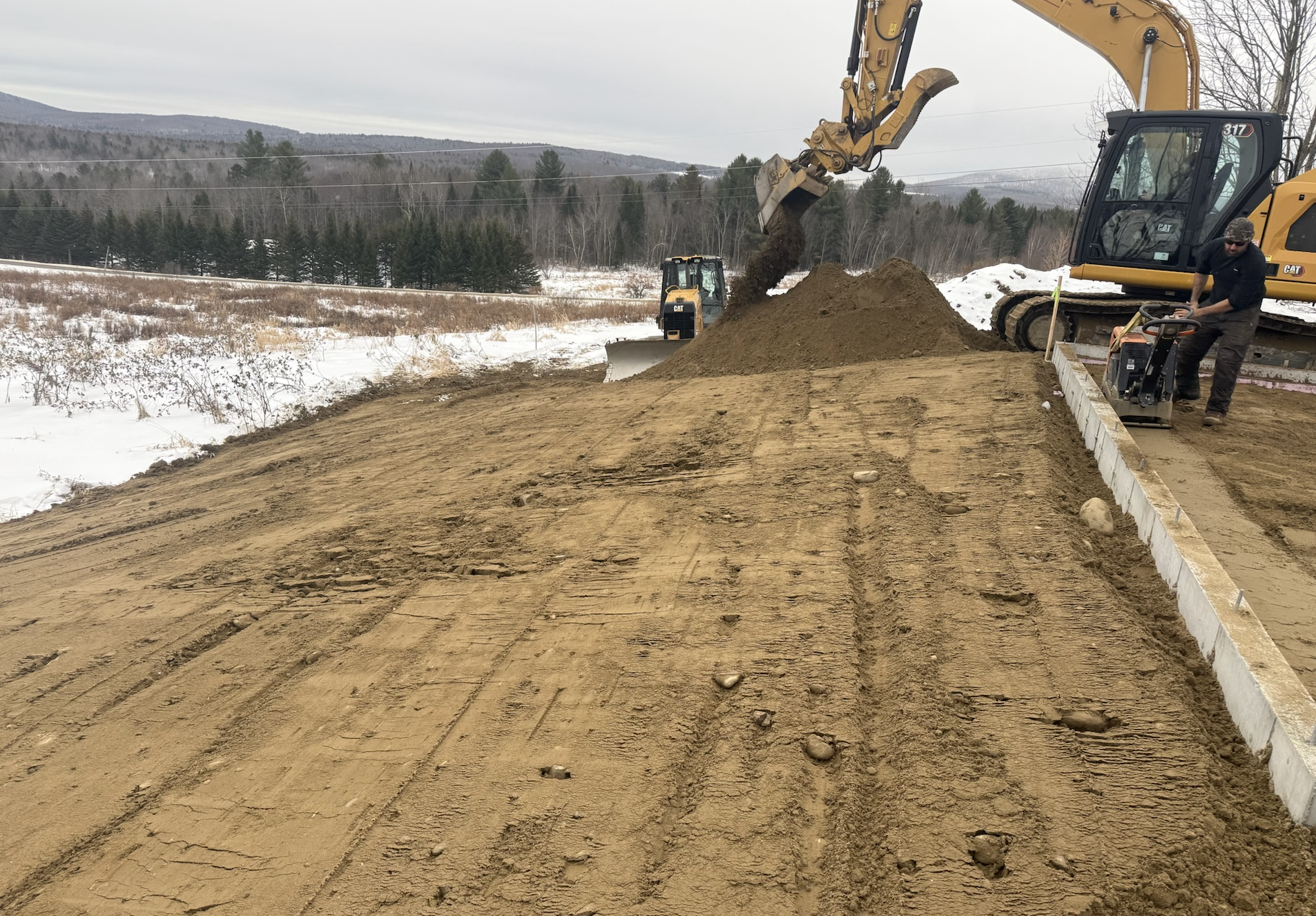 Construction site with yellow excavators and a worker using a compactor, building a road in a snowy, rural landscape with trees and hills in the background.