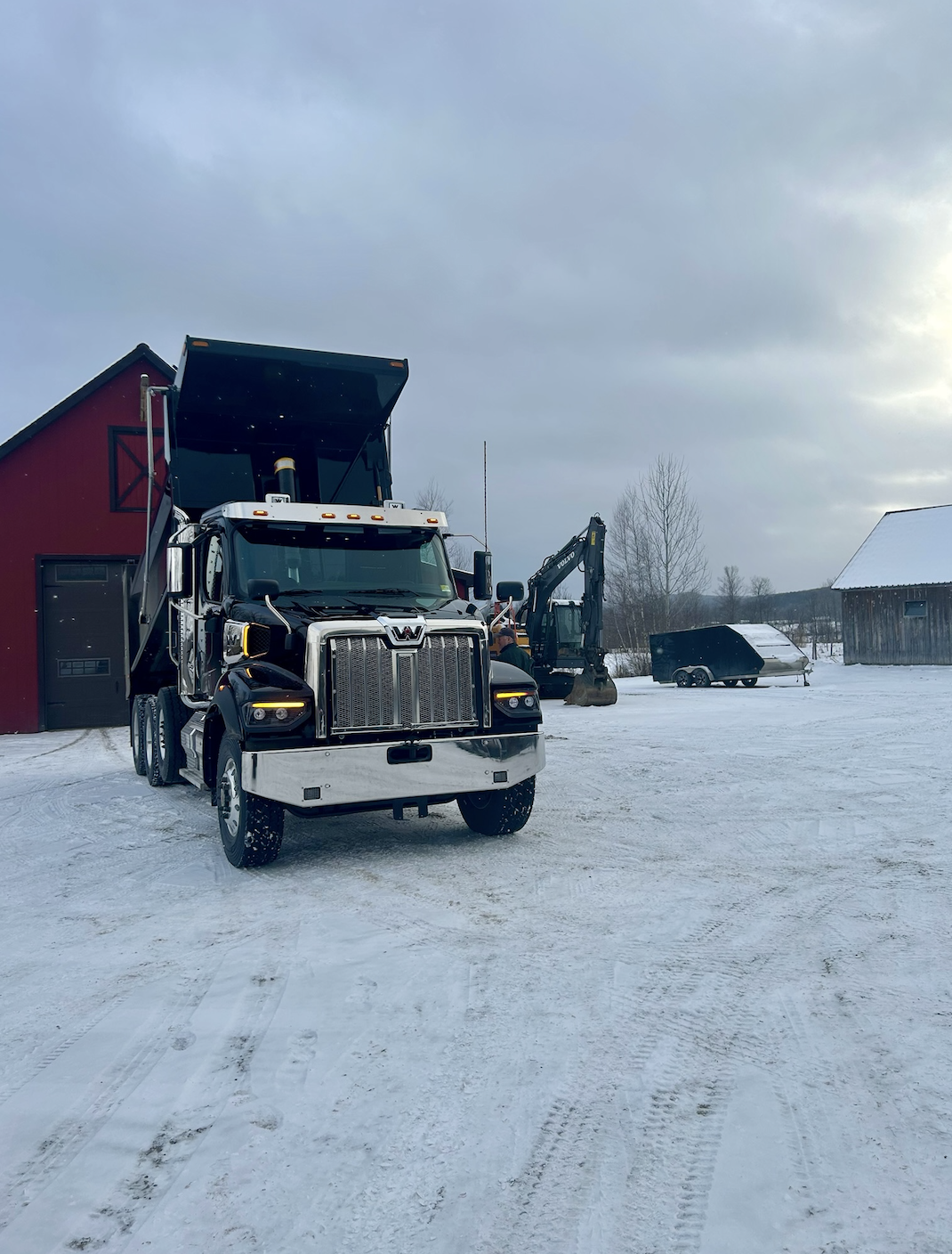 A large black and chrome semi-truck parked on a snow-covered farm or yard, with a red barn and a wood building in the background, along with a small excavator and a trailer.