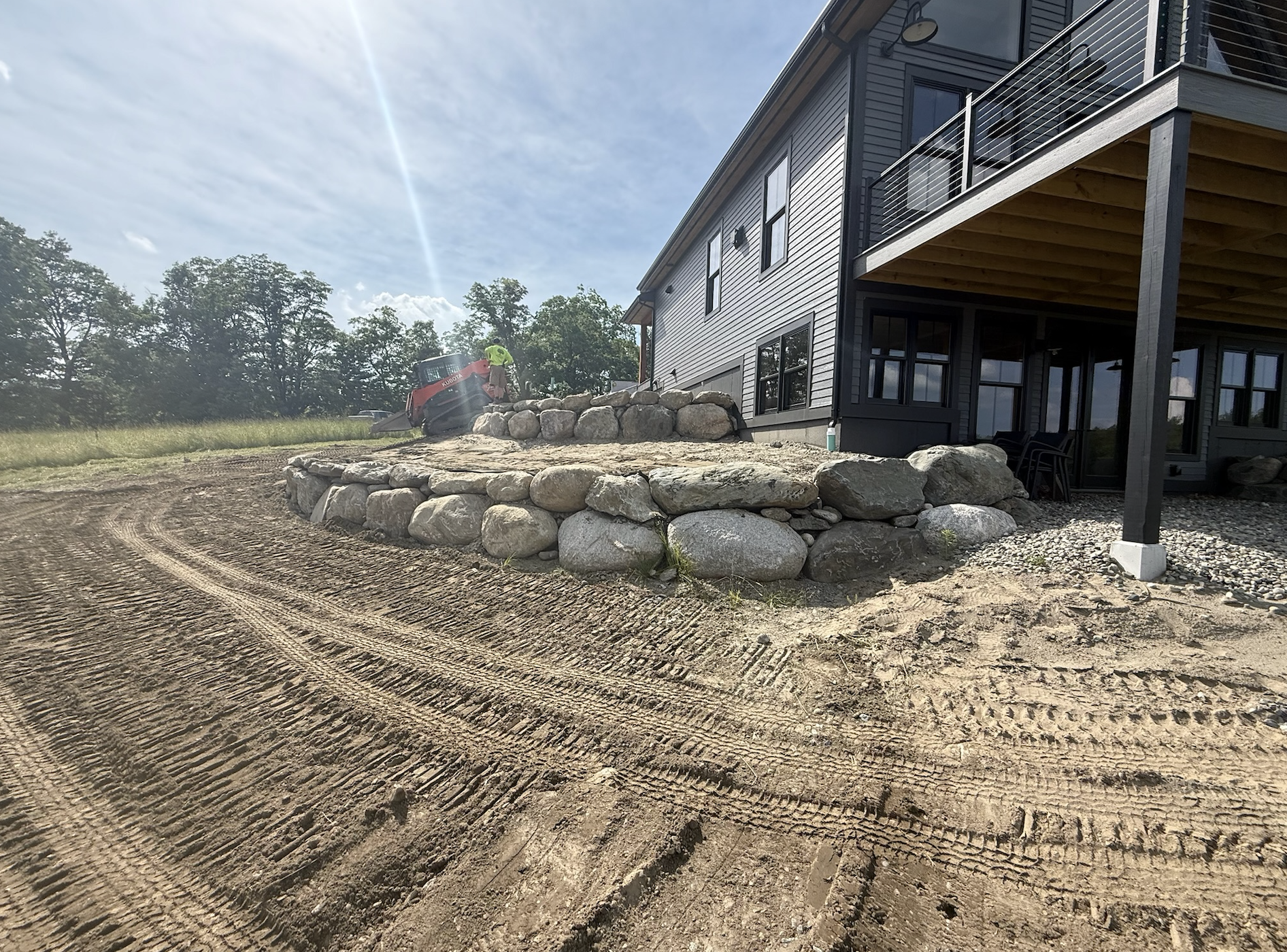 A backyard landscape under construction with a stone retaining wall, a dirt driveway, and a modern house with large windows and a balcony. A worker is operating a compact excavator.