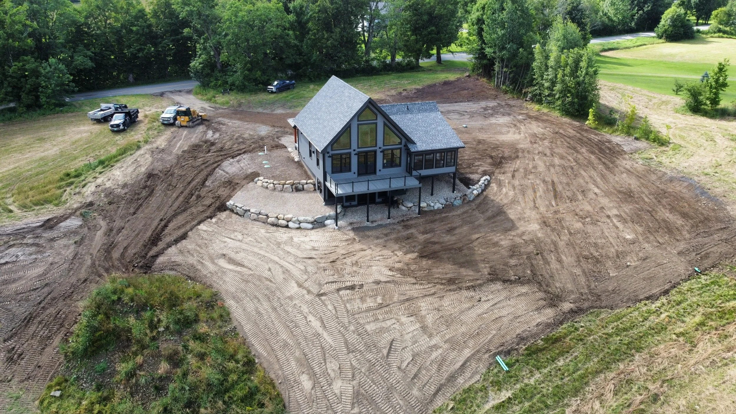 A new house under construction in a cleared plot of land with tire marks on the dirt, surrounded by trees and some parked vehicles.