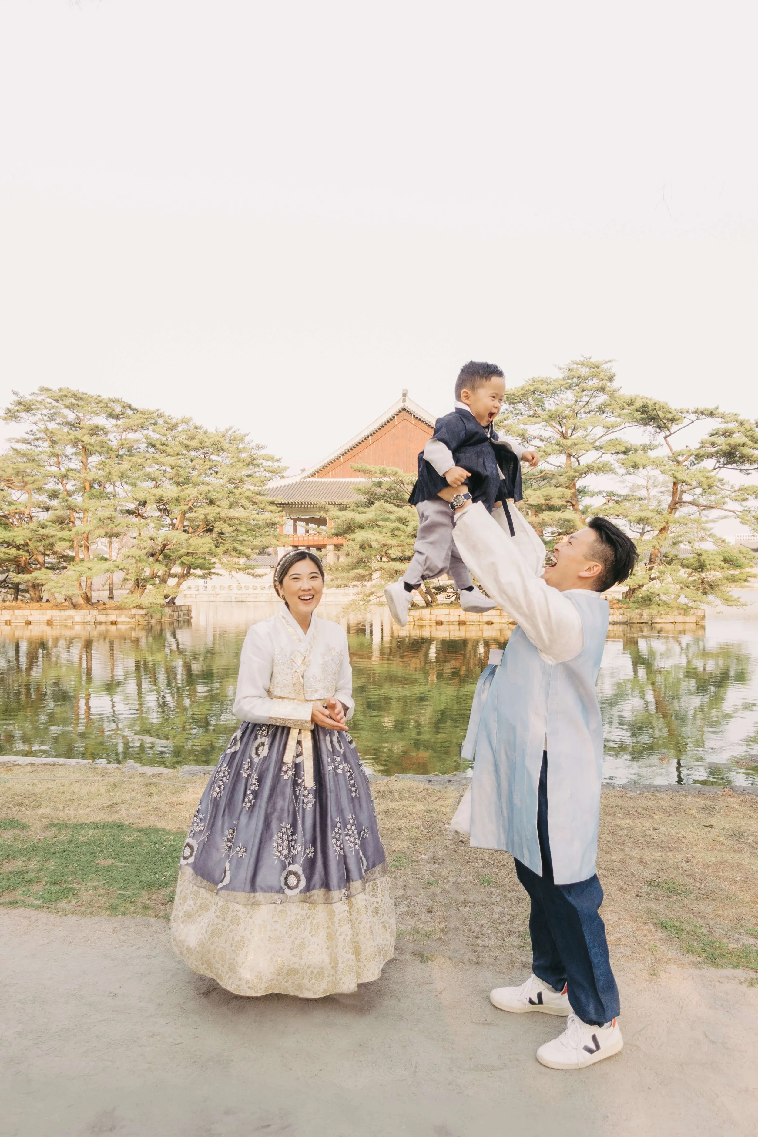 Family wearing traditional Korean hanbok near a pond, with trees and a traditional building in the background. Man lifting child into the air while woman smiles.