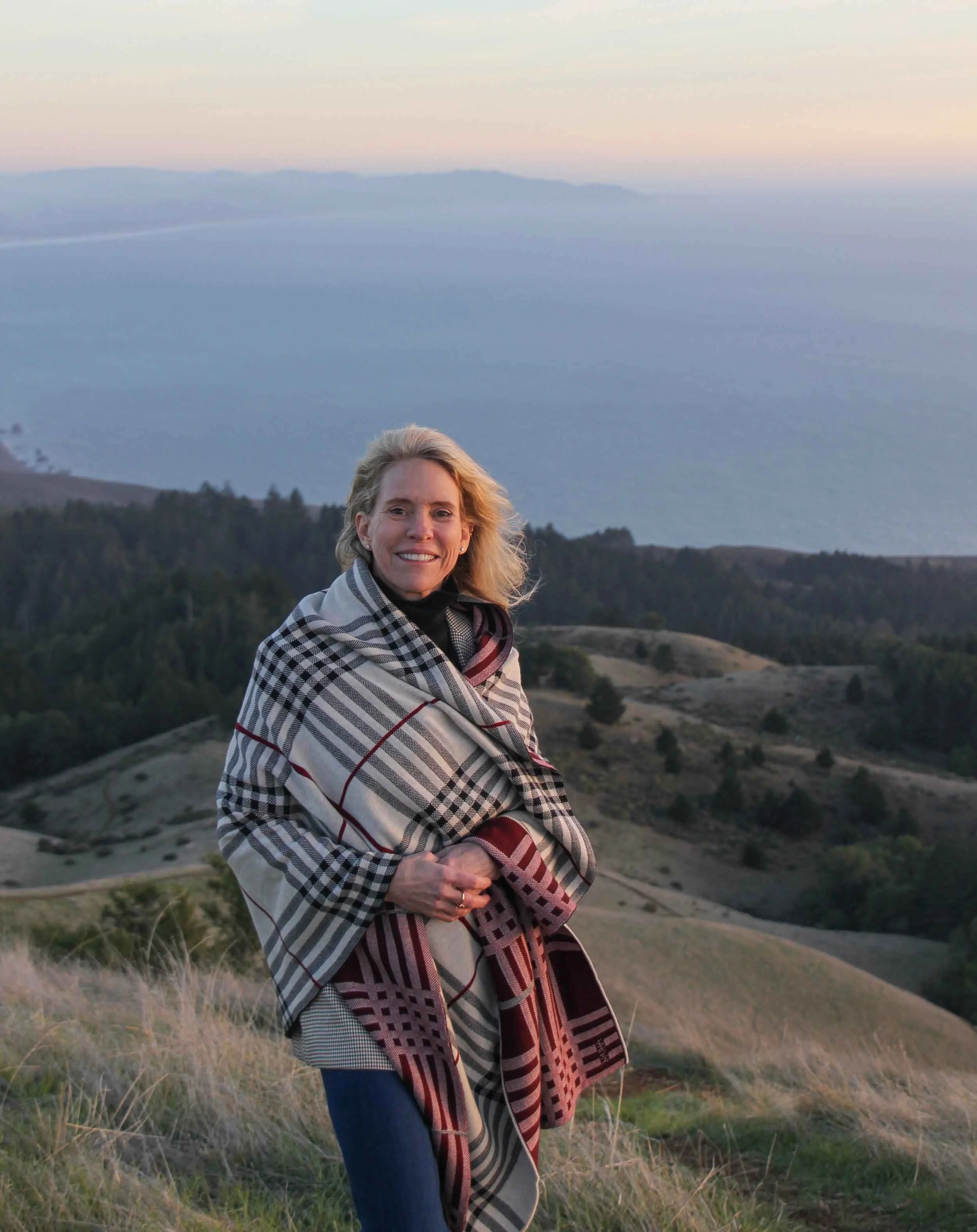 A woman standing outdoors on a hill at sunset, wearing a black turtleneck and wrapping herself in a patterned blanket with hills and water in the background.