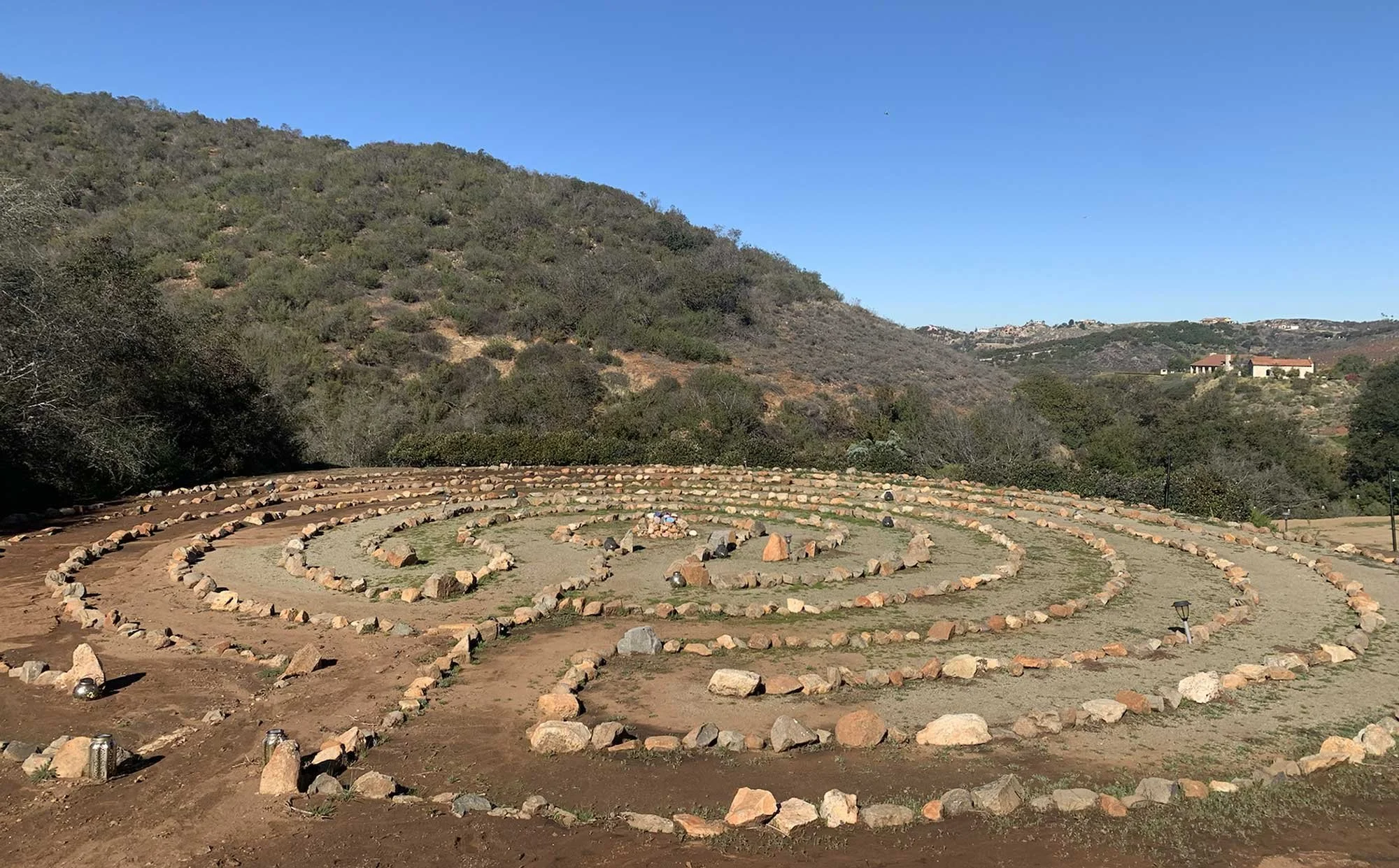 A spiral labyrinth made of rocks on a dirt ground with distant hills and houses under a clear blue sky.