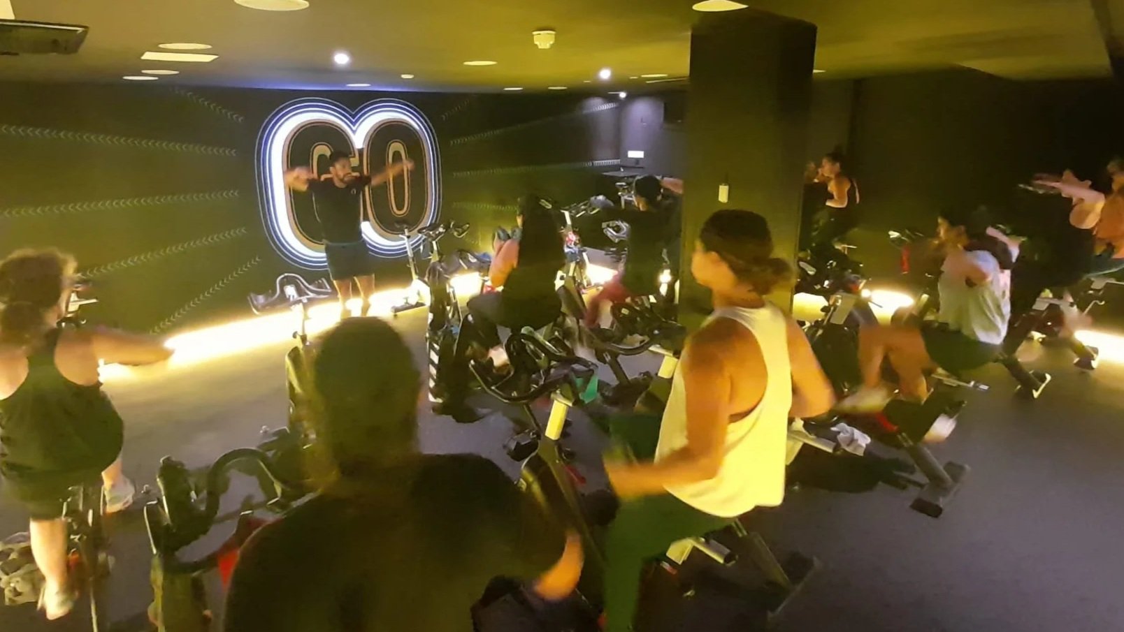 Indoor cycling class with people on stationary bikes facing a instructor at the front, with a large illuminated 'GO' sign on the dark wall.
