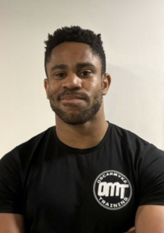 A young man with short curly hair and a beard, wearing a black t-shirt with 'OCARMY FITNESS' and 'OMT' logos, standing against a plain beige wall.