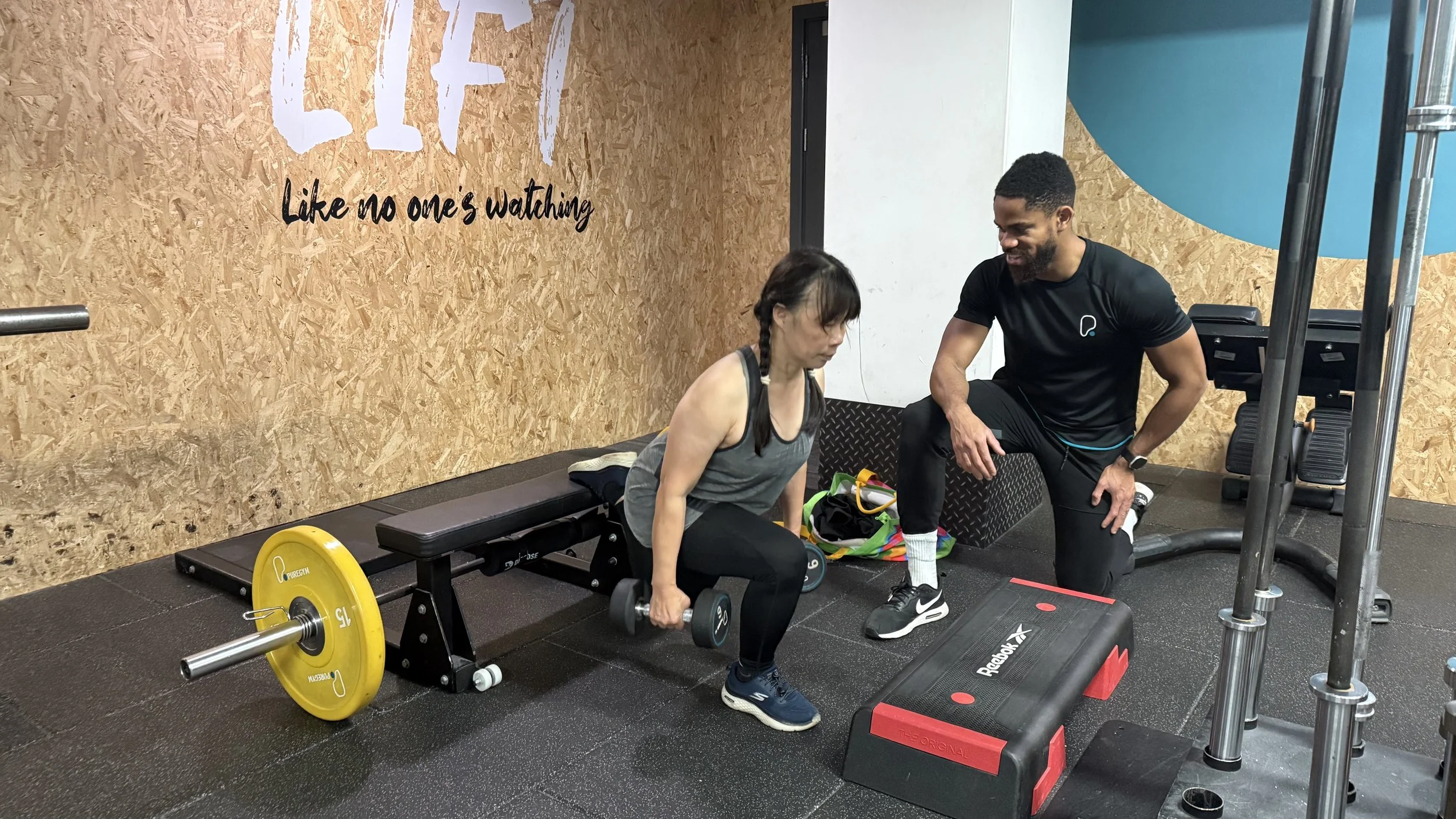 A woman performs a deadlift exercise with a barbell at a gym while a male trainer watches and encourages her.