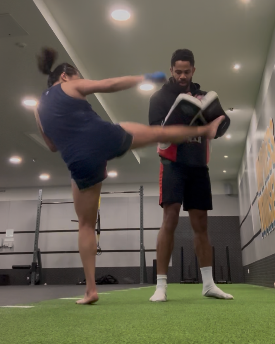 A woman performing a high kick towards a pad held by a man during a martial arts training session in a gym.