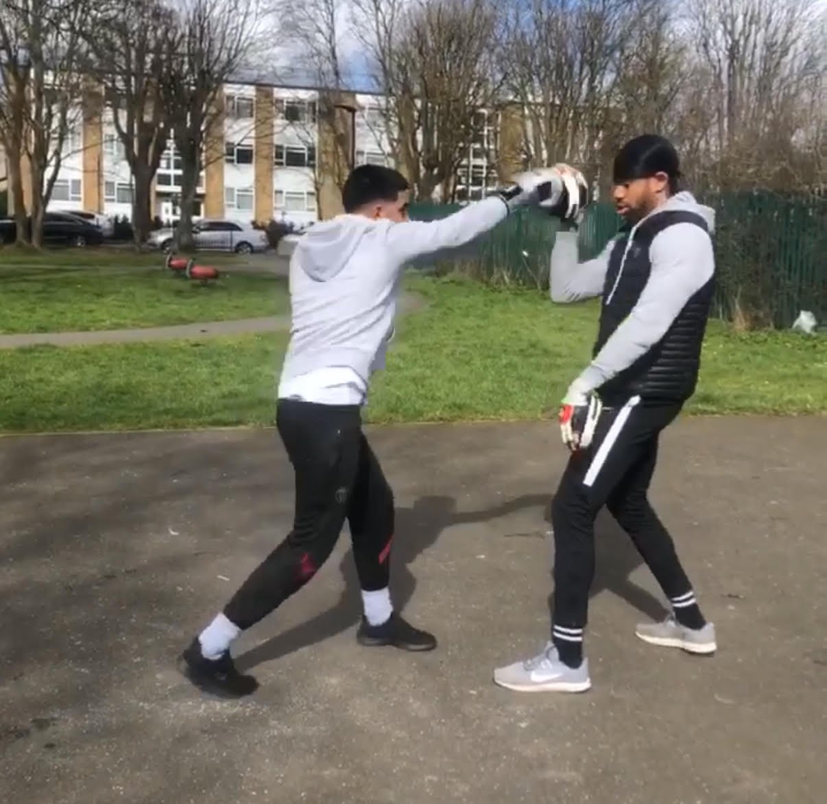 Two young men practicing boxing outdoors, one throwing a punch while the other is in a defensive stance, on a paved area with grass and trees in the background.
