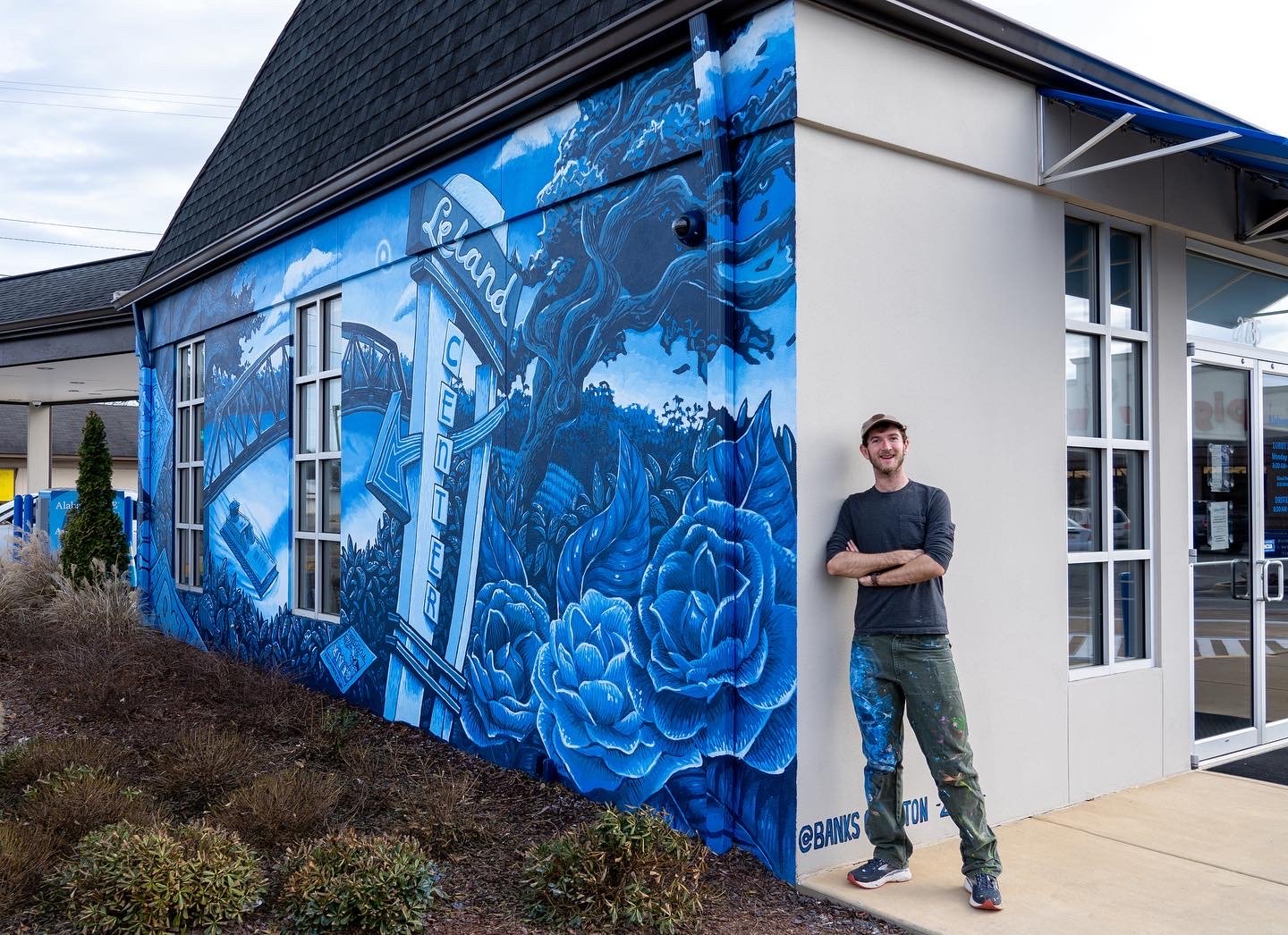 A man standing outside a building with blue-themed mural art featuring a tree, flowers, and the letter C en route to a downtown area.