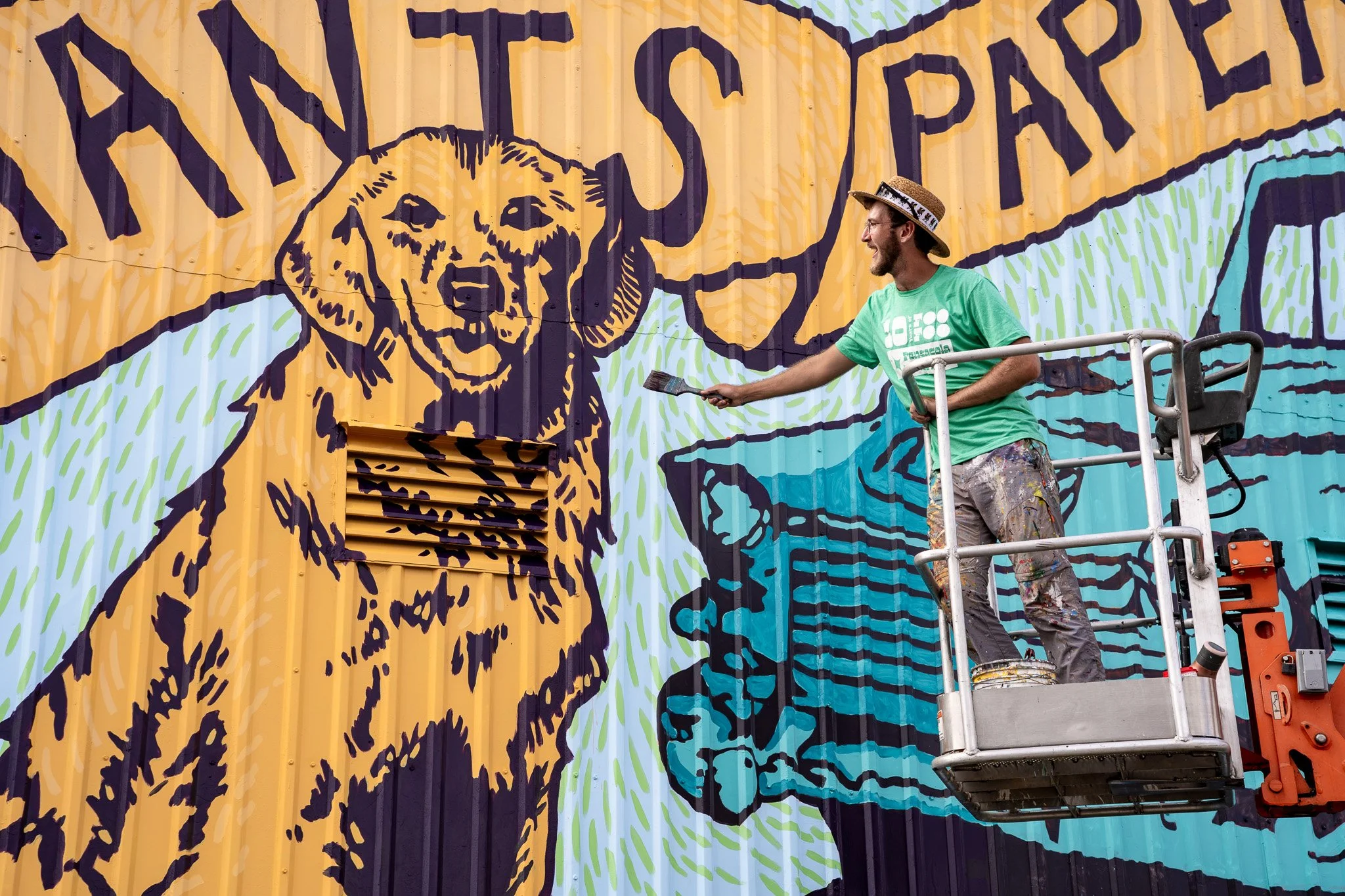 Banks Compton painting a large commercial mural from a lift at Merchants Paper Company in Pensacola, Florida.