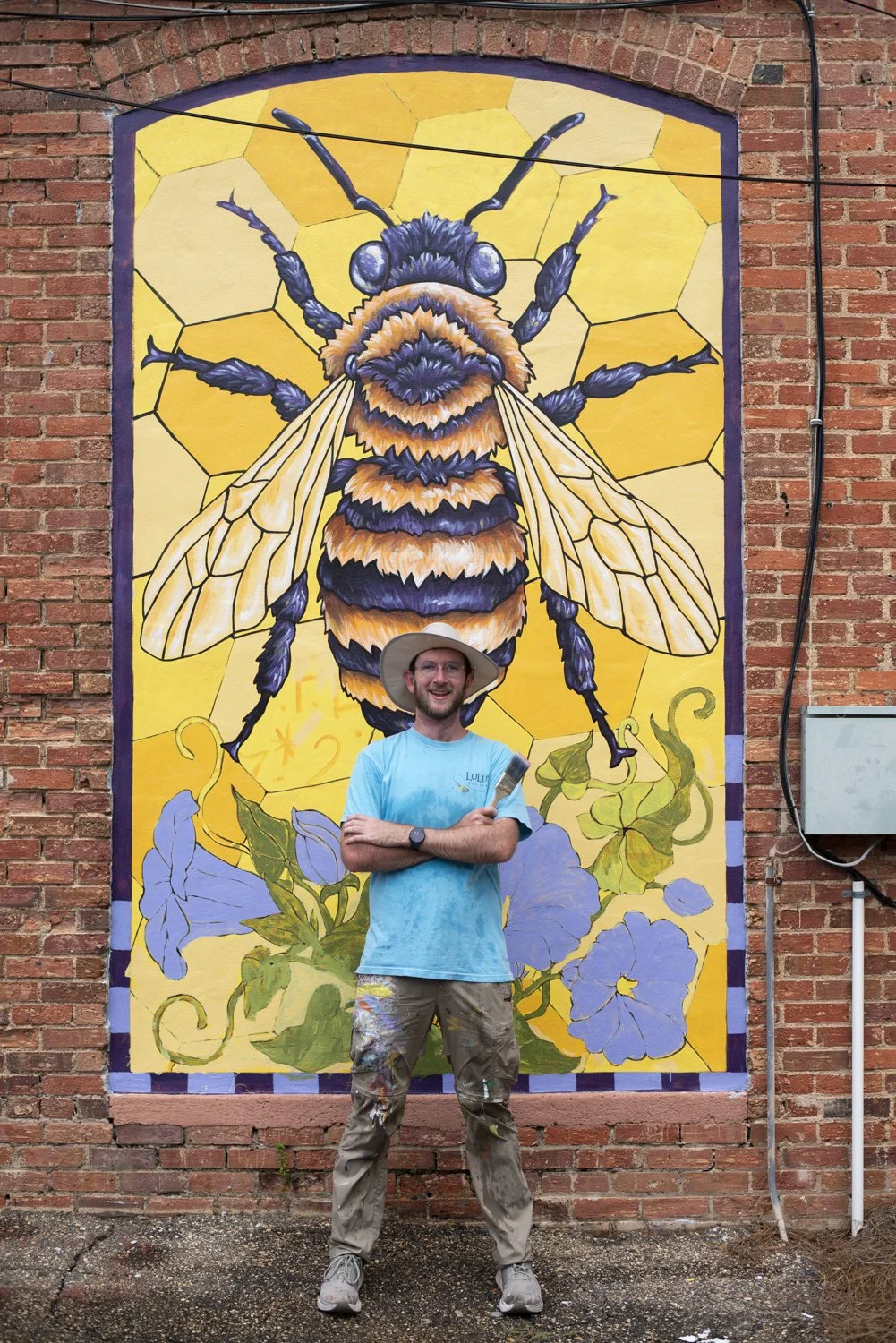 A man standing with arms crossed, smiling, in front of a large vibrant mural of a bee on a brick wall. The man is wearing a light blue shirt, paint-stained pants, and a wide-brimmed hat.
