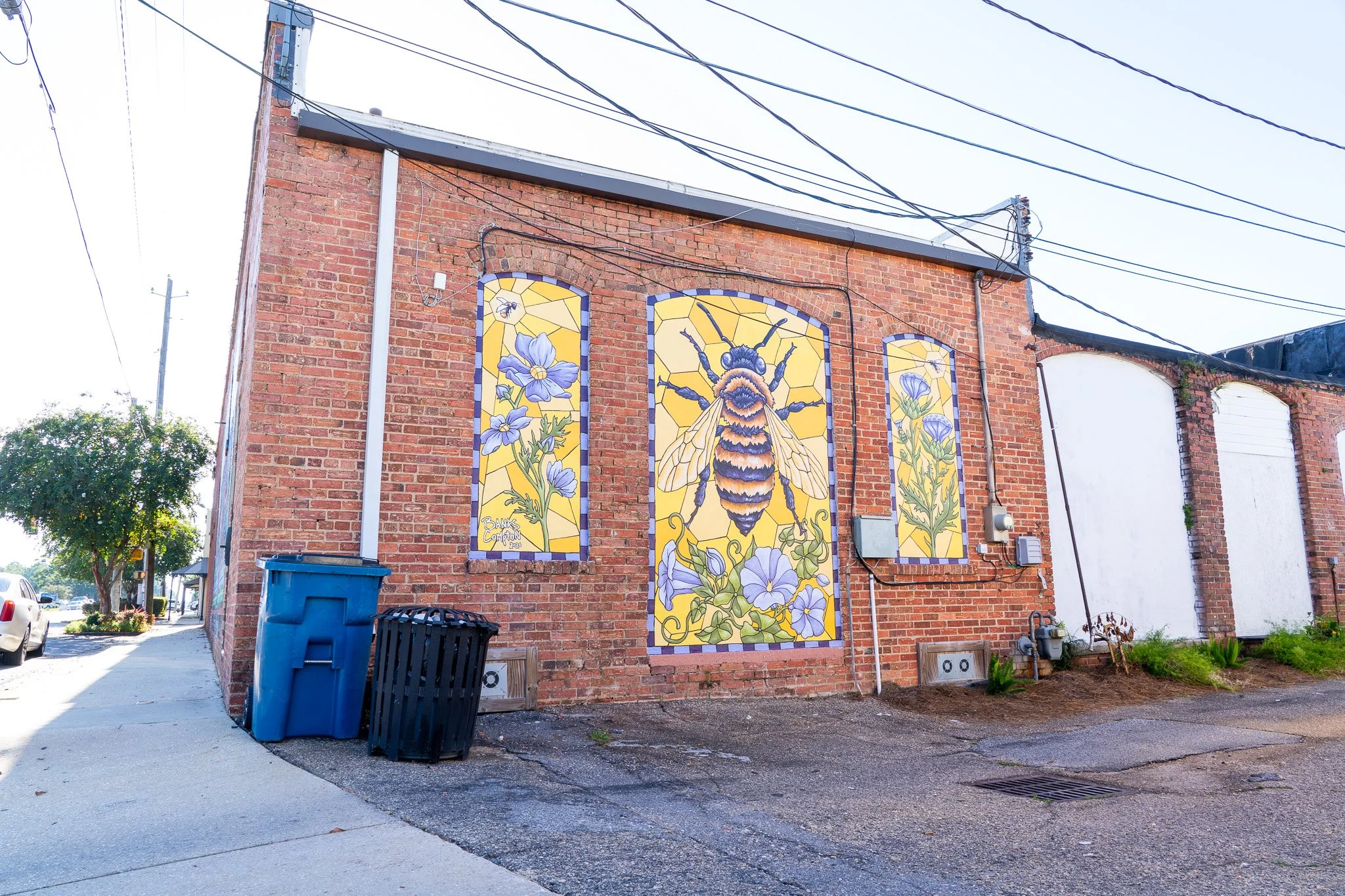Mural on brick building depicting a large bee surrounded by purple flowers with a yellow background.