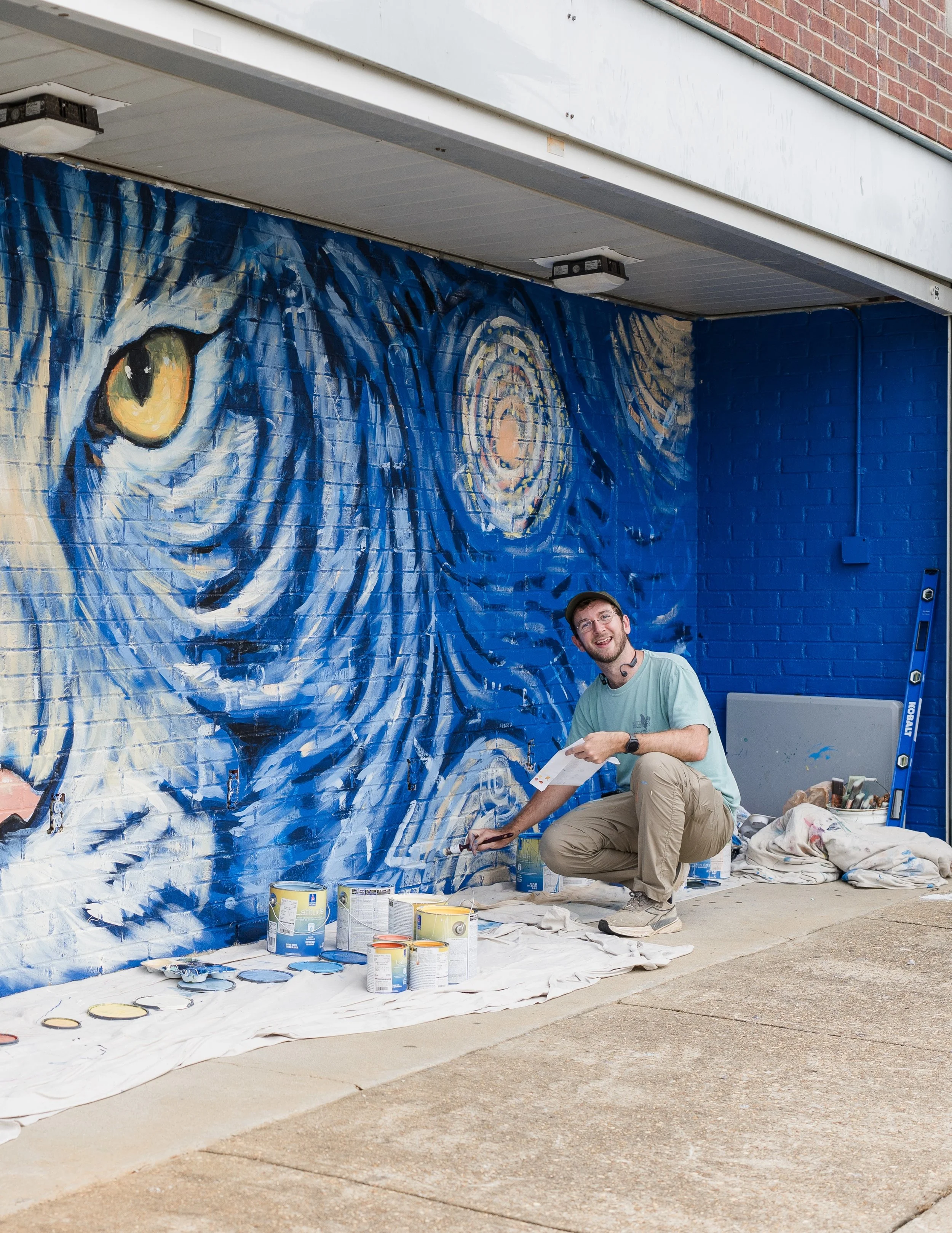 A man squatting in front of a large wall mural of an eye painted on a brick wall, surrounded by cans of paint, brushes, and painting supplies outdoors.