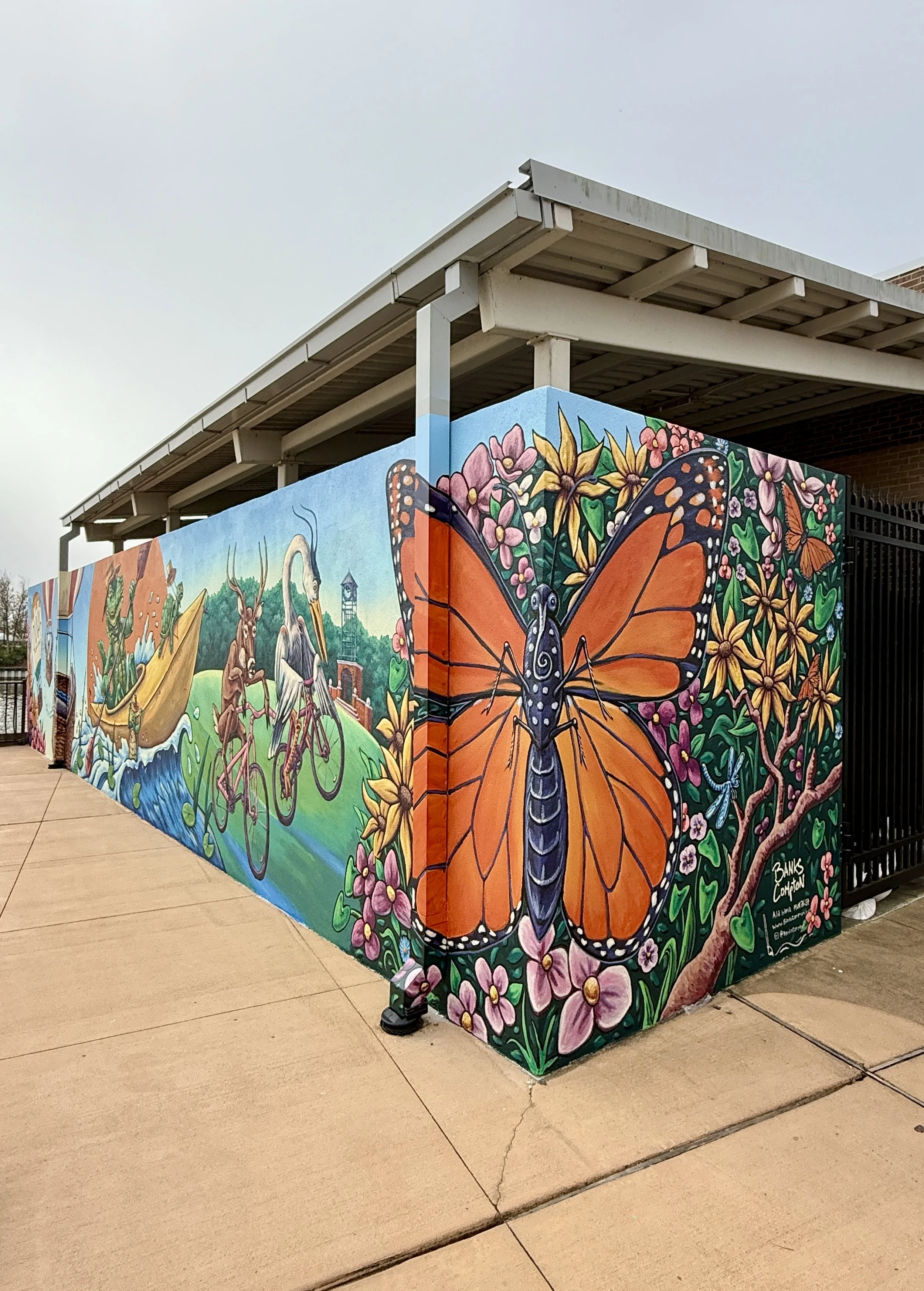 Colorful mural art featuring a large butterfly with orange and black wings, surrounded by flowers, trees, and various animals on an outdoor wall.