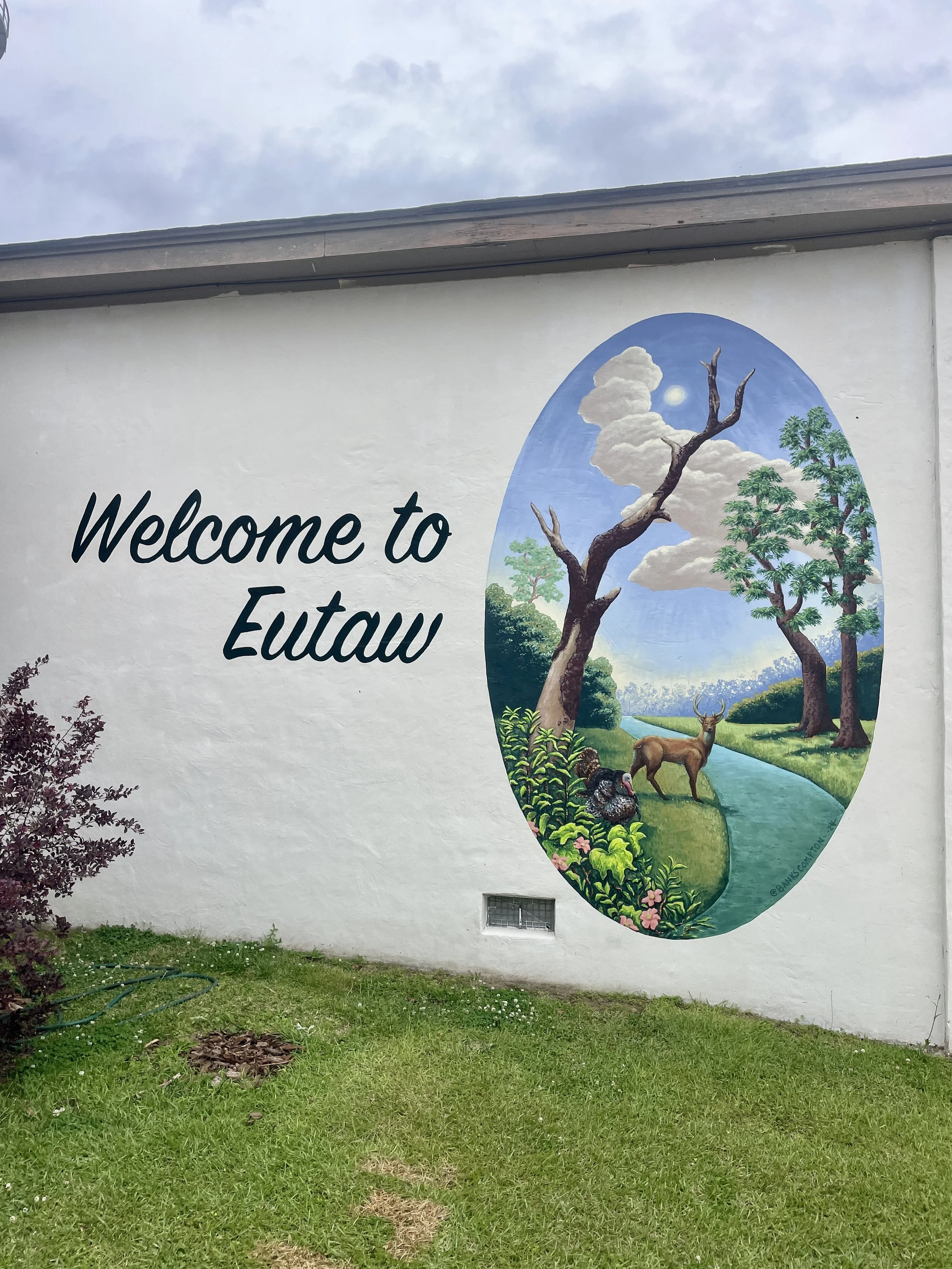 Welcome to Eutaw mural by Banks Compton in Eutaw, Alabama, featuring a white-tailed deer, wild turkey, water, trees, and West Alabama landscape.