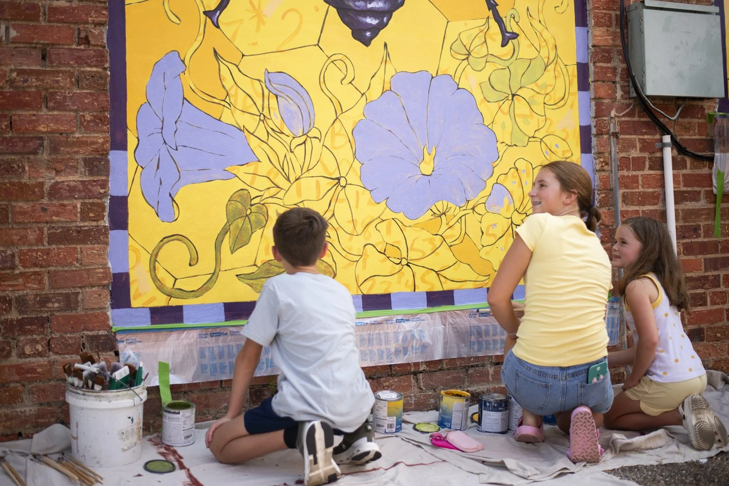 Children and a woman painting a large mural of blue and yellow flowers on a brick wall.