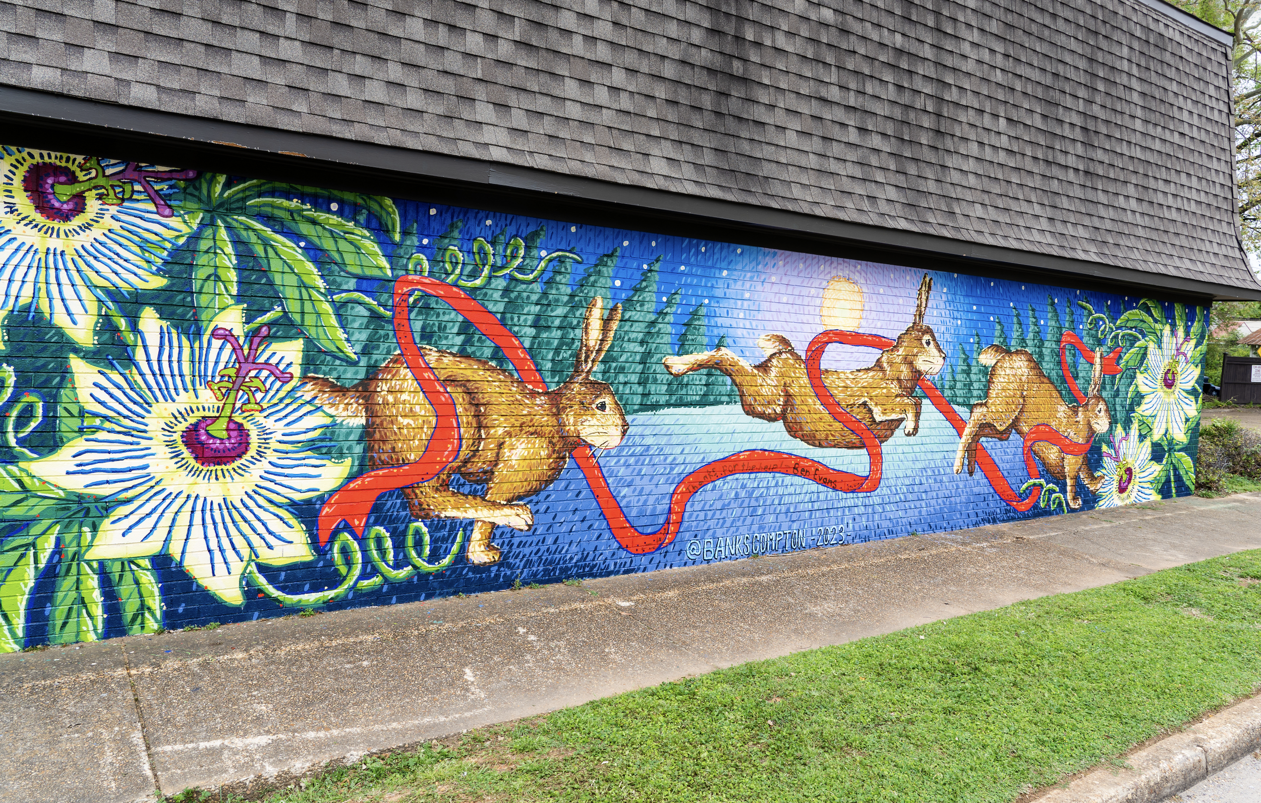 Colorful mural painted on a brick wall featuring rabbits, bright flowers, and a red snake, with a dark roof above and green grass below.