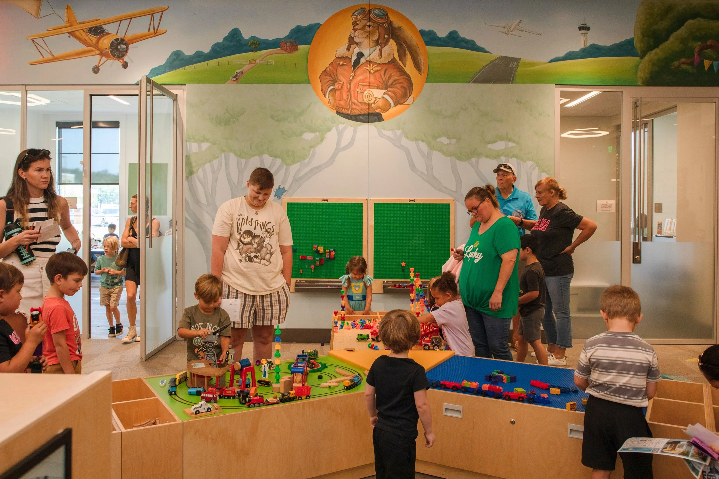 Children and families inside Foley Community Library beneath Banks Compton's storybook mural featuring local aviation imagery and whimsical animal characters.