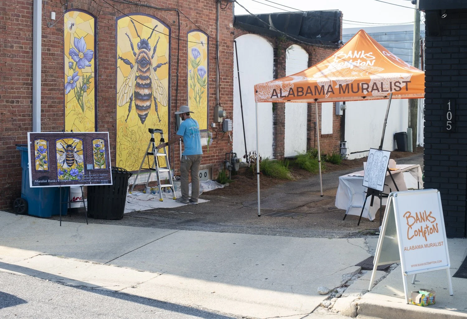An artist painting a mural of a bee on a brick wall, with a sign and tent displaying the words 'Alabama Muralist' and 'Banks Compton'.