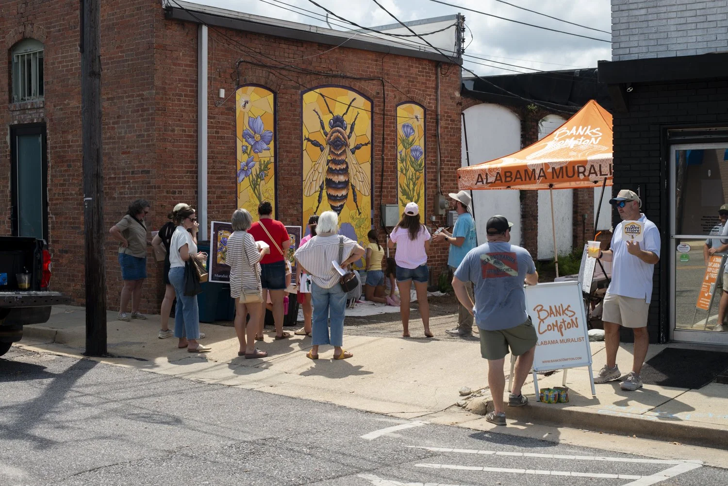 A group of people gathered outside a brick building with a colorful mural of a bee and purple flowers. There is a white tent with orange lettering reading 'ALABAMA MURALIST' and a sign for 'Banks Compton,' indicating an event related to mural art. Some people are looking at artwork or taking photos.