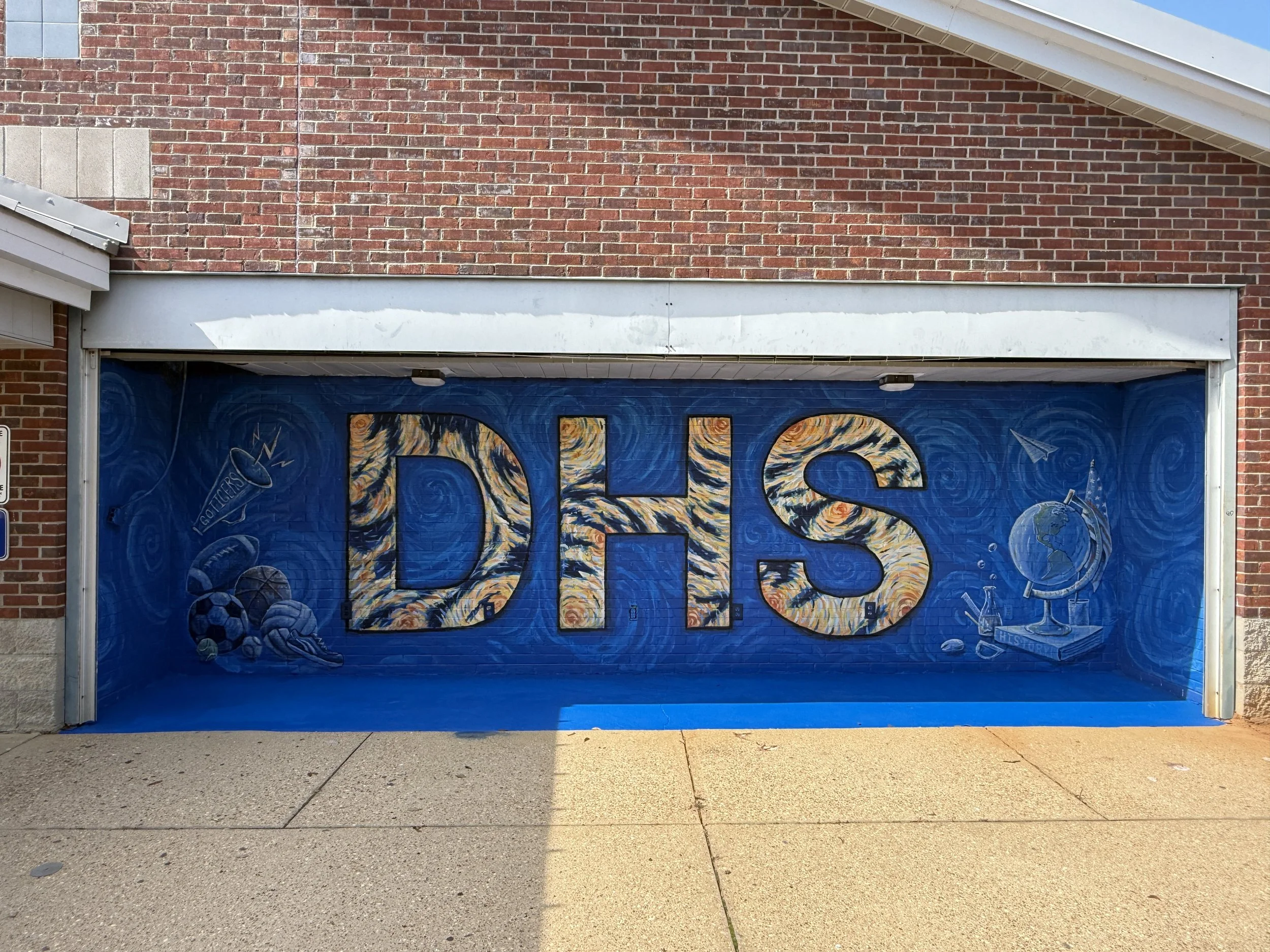 A blue garage door with the word 'DHS' painted on it in large, colorful, signature-style letters. The garage is attached to a brick building with a sloped roof, and the ground in front is a concrete sidewalk.
