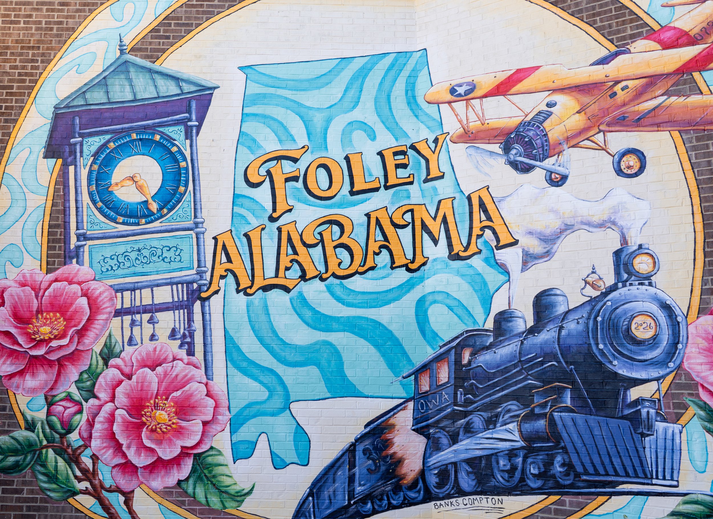 Foley Photo Mural by Alabama mural artist Banks Compton at Downtown OWA, featuring the Heritage Park clock tower, Foley Alabama lettering, historic train, N3N biplane, and camellias.