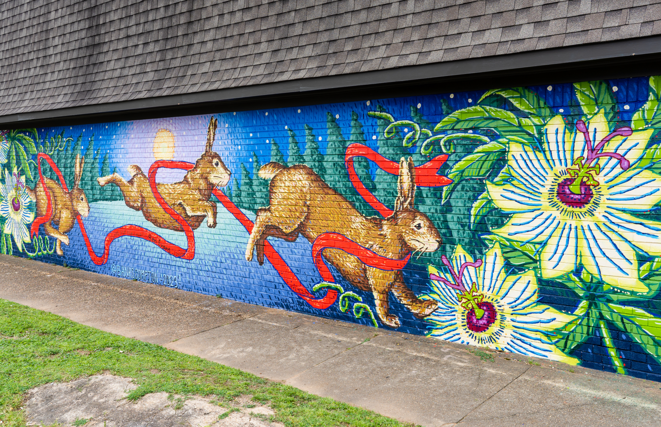 Colorful mural of animals and flowers painted on a brick wall with a gray roof above and grass below.
