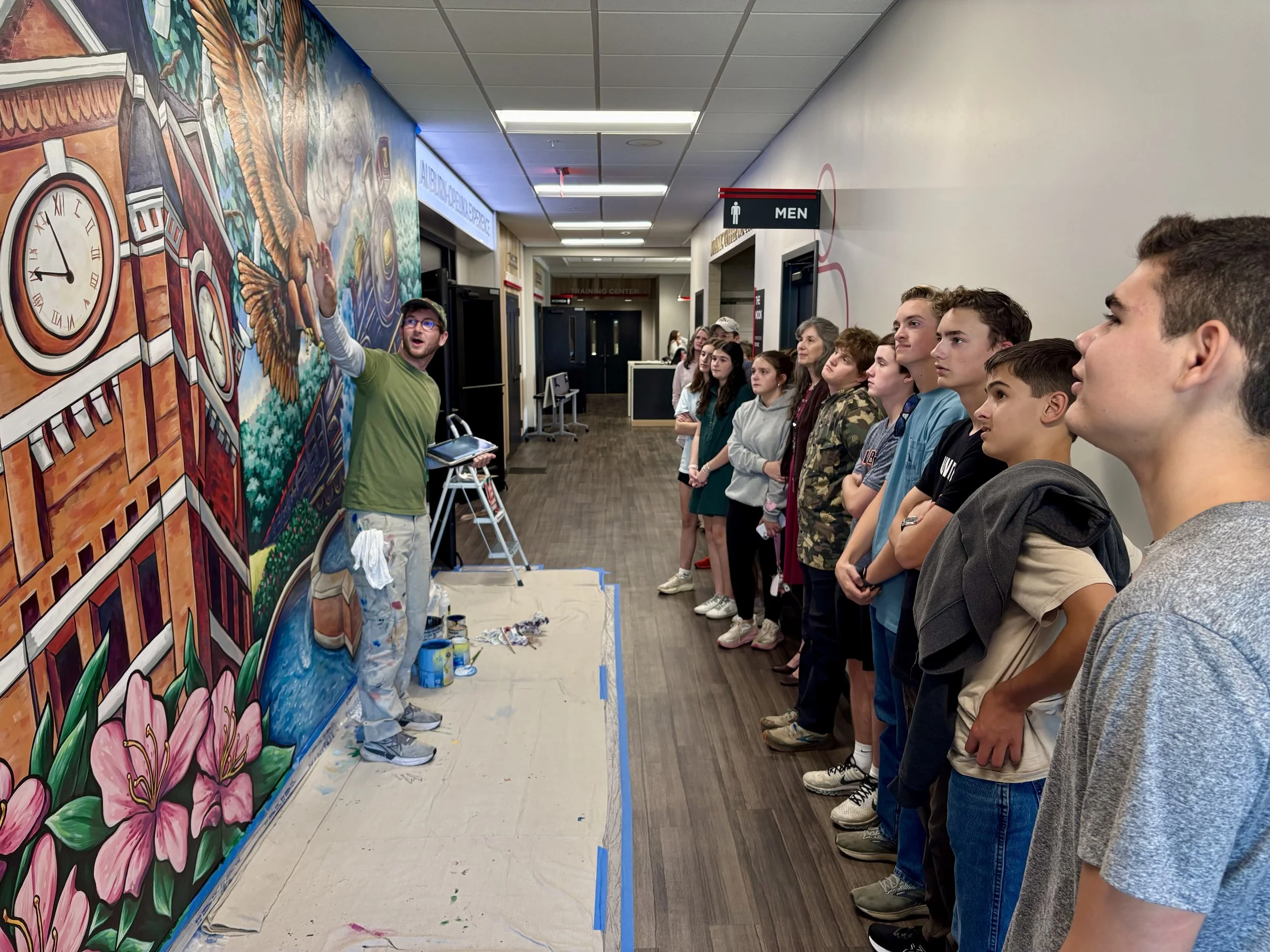 A man in a green shirt and glasses is painting a colorful mural on a wall while a group of young people watch. The mural features flowers, an owl, a clock, and other vibrant elements. The scene takes place in an indoor hallway with signs indicating restrooms and a training center.
