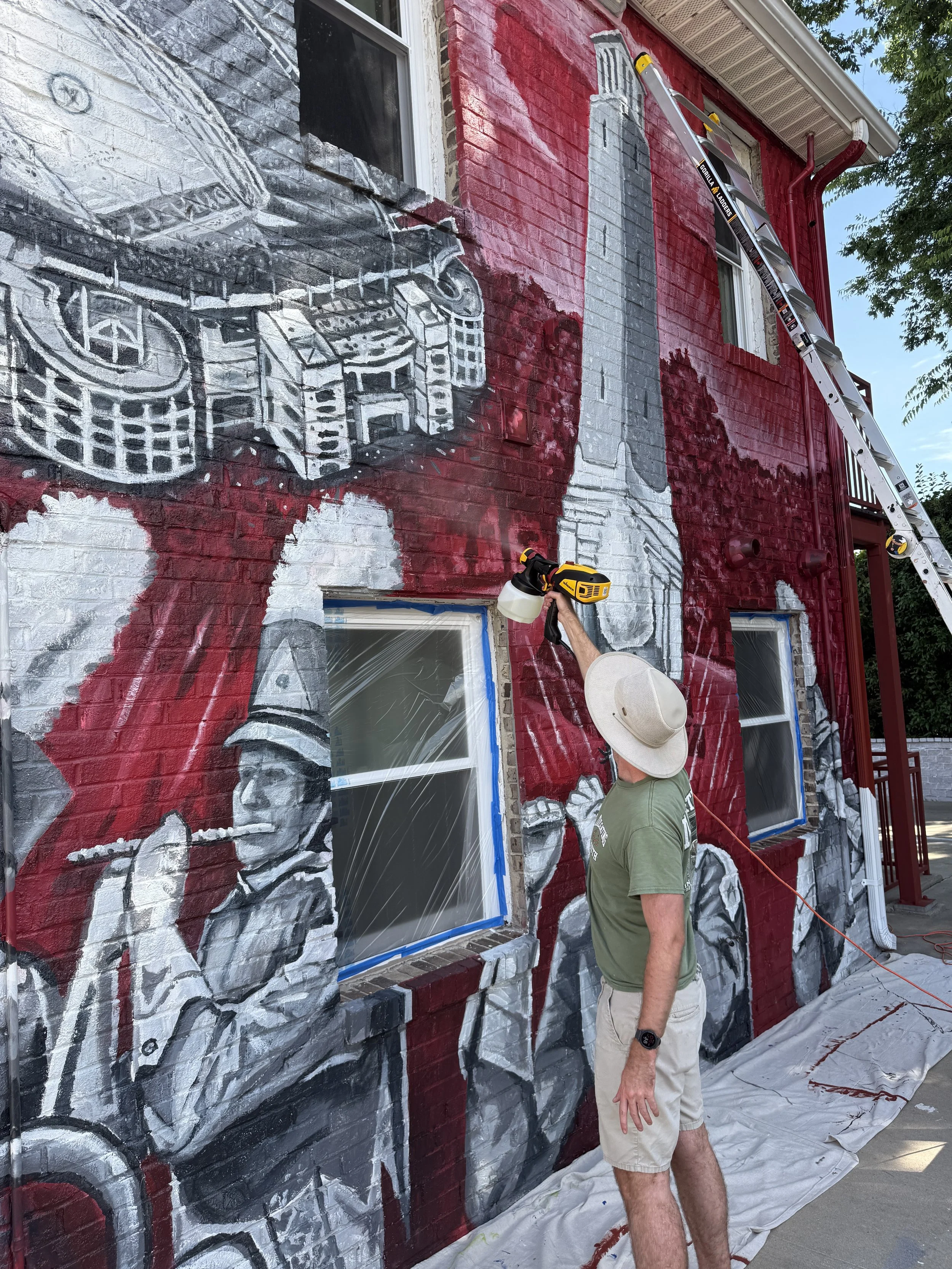 A person painting a mural on a brick building, showing a partial black and white design next to a large gray and white rocket with a red background. The person is wearing a hat and holding a paint sprayer, with a ladder leaning against the building.
