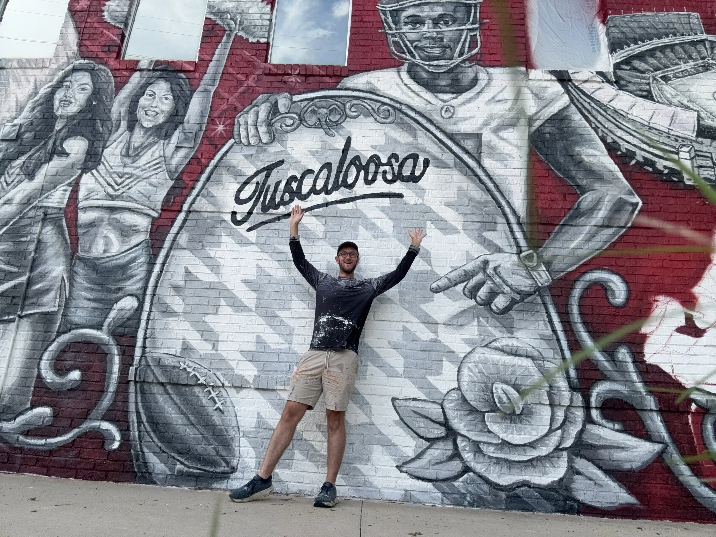 A man standing in front of a large mural on a brick wall, raising the arms with palms facing outward. The mural is in black, white, and red and depicts two women and a young man wearing a football helmet. The mural features the word 'Consejo' written in script.
