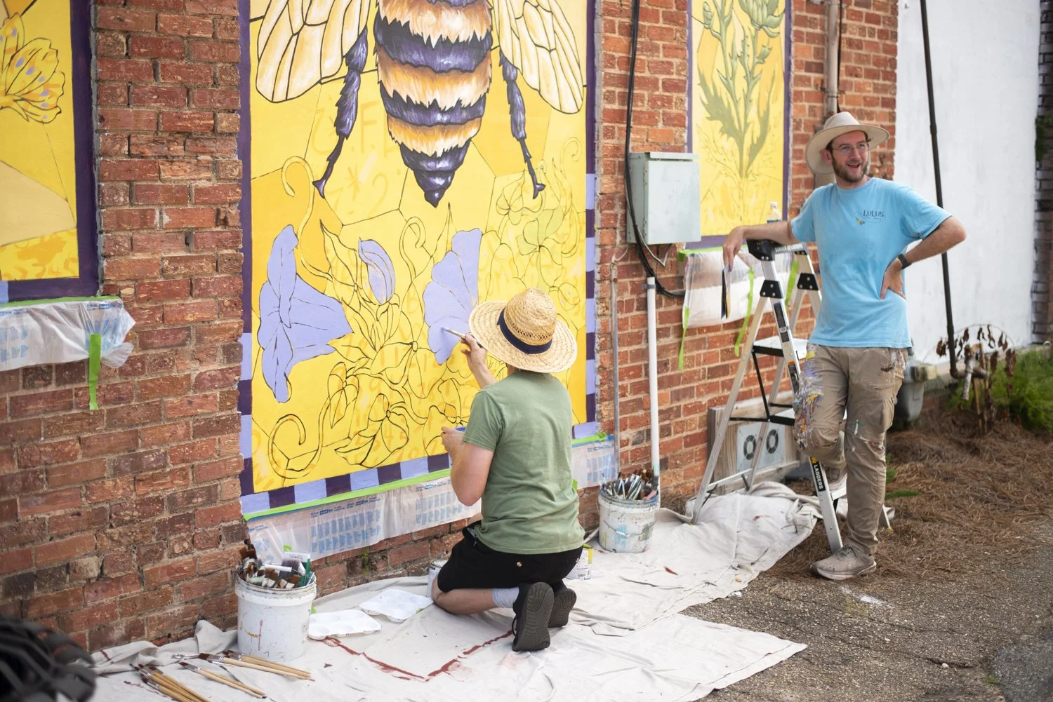 Two people painting a large mural of a bee with yellow and lavender flowers on a brick wall outdoors. One person is kneeling and wearing a straw hat, the other is standing wearing a wide-brimmed hat and glasses.