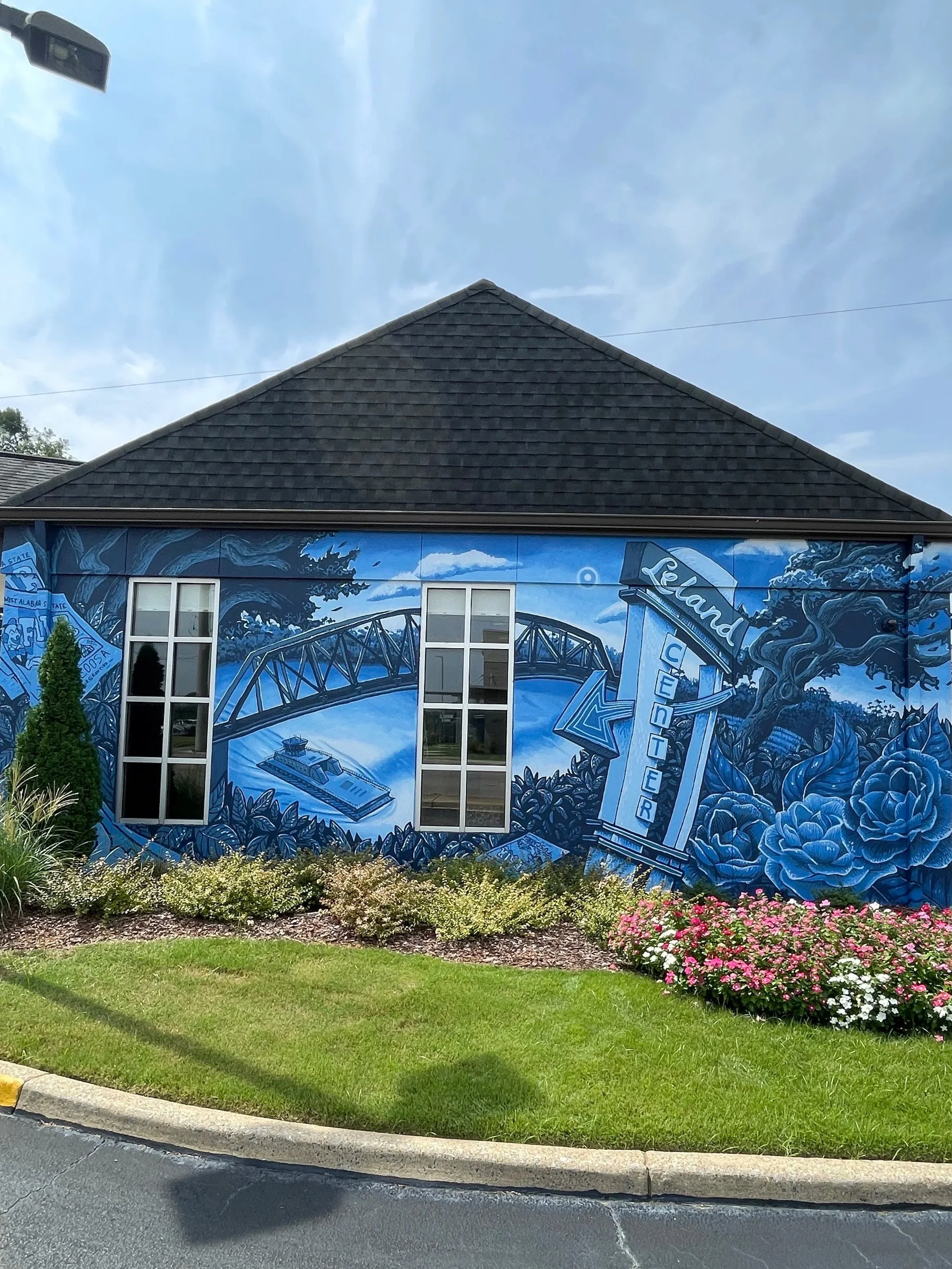 A large blue mural on the side of a building depicting a roller coaster, a boat, and a bottle of water, with the words "Land Center." There are two windows in the mural, and a small garden with various flowers and bushes in front of the building. The sky is partly cloudy.