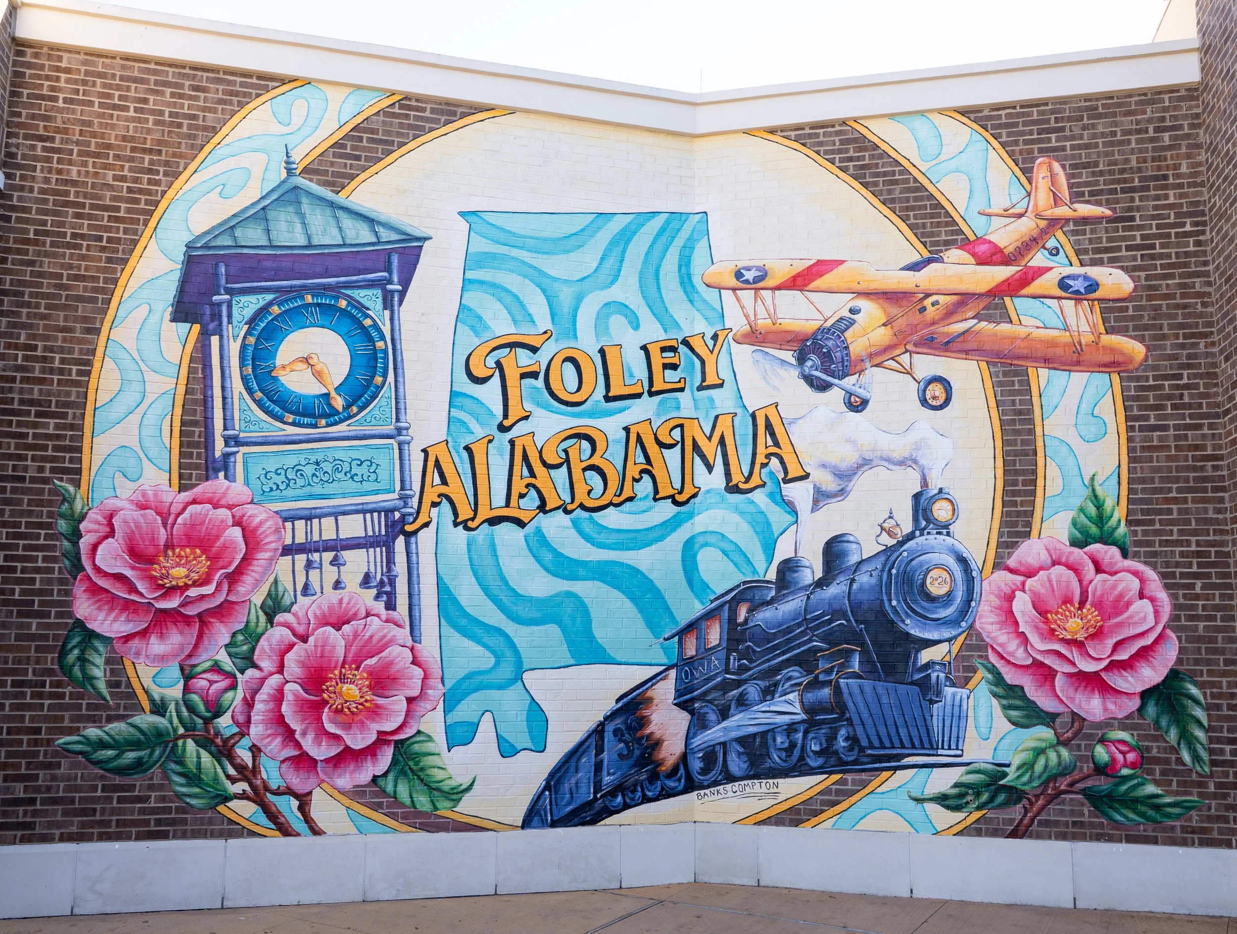 OWA Foley Alabama mural by Banks Compton featuring the Heritage Park clock tower, a historic train, N3N biplane, camellias, and large Foley Alabama lettering.