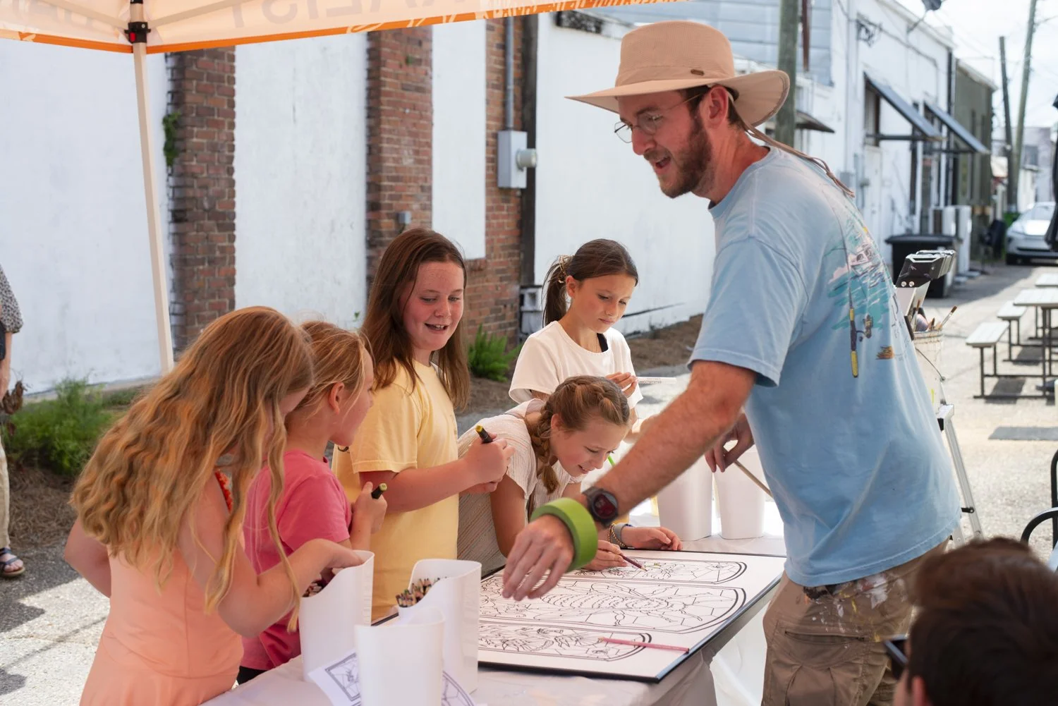 An outdoor art activity with children at a booth, where an adult is guiding them in drawing or coloring. The setting appears to be on a sidewalk near brick and white buildings, with tables, chairs, and art supplies visible.