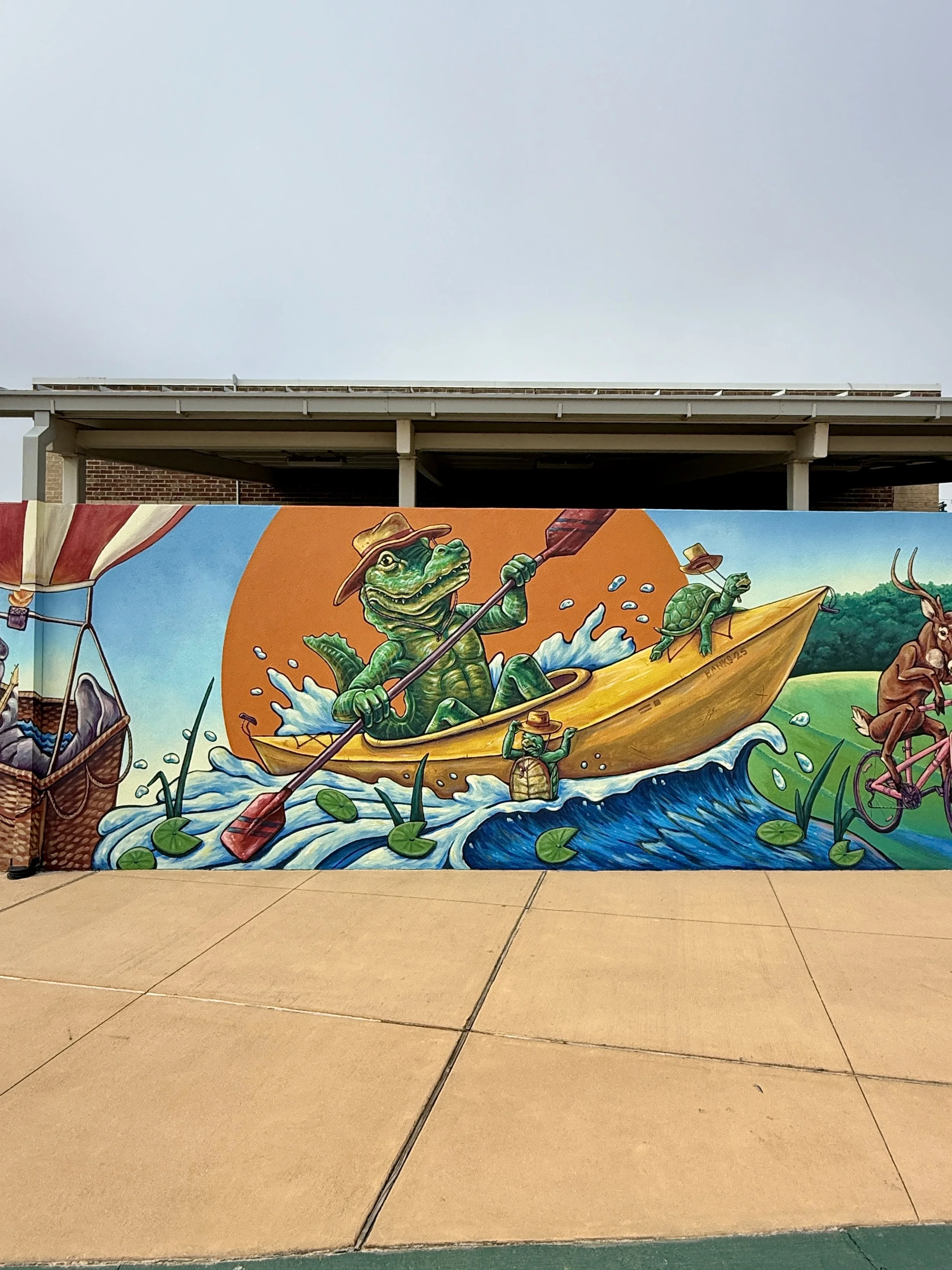 Colorful mural of frogs kayaking on water, with water lilies and aquatic plants, in front of a building with a gray sky overhead.