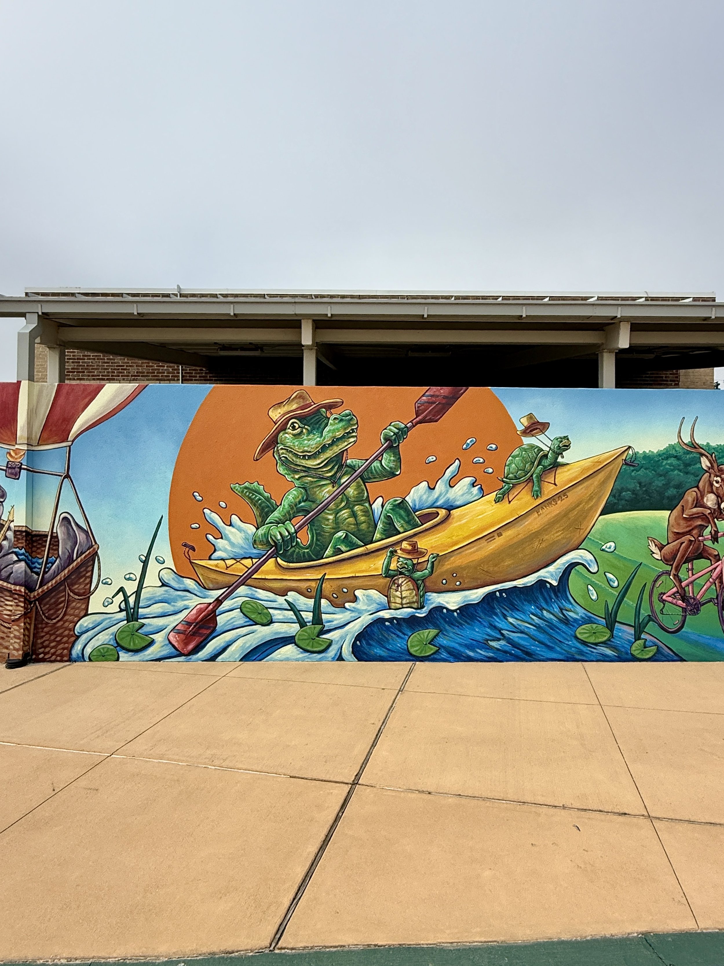Downtown OWA mural by Banks Compton in Foley, Alabama, featuring an alligator and turtle kayaking through a playful Gulf Coast landscape.