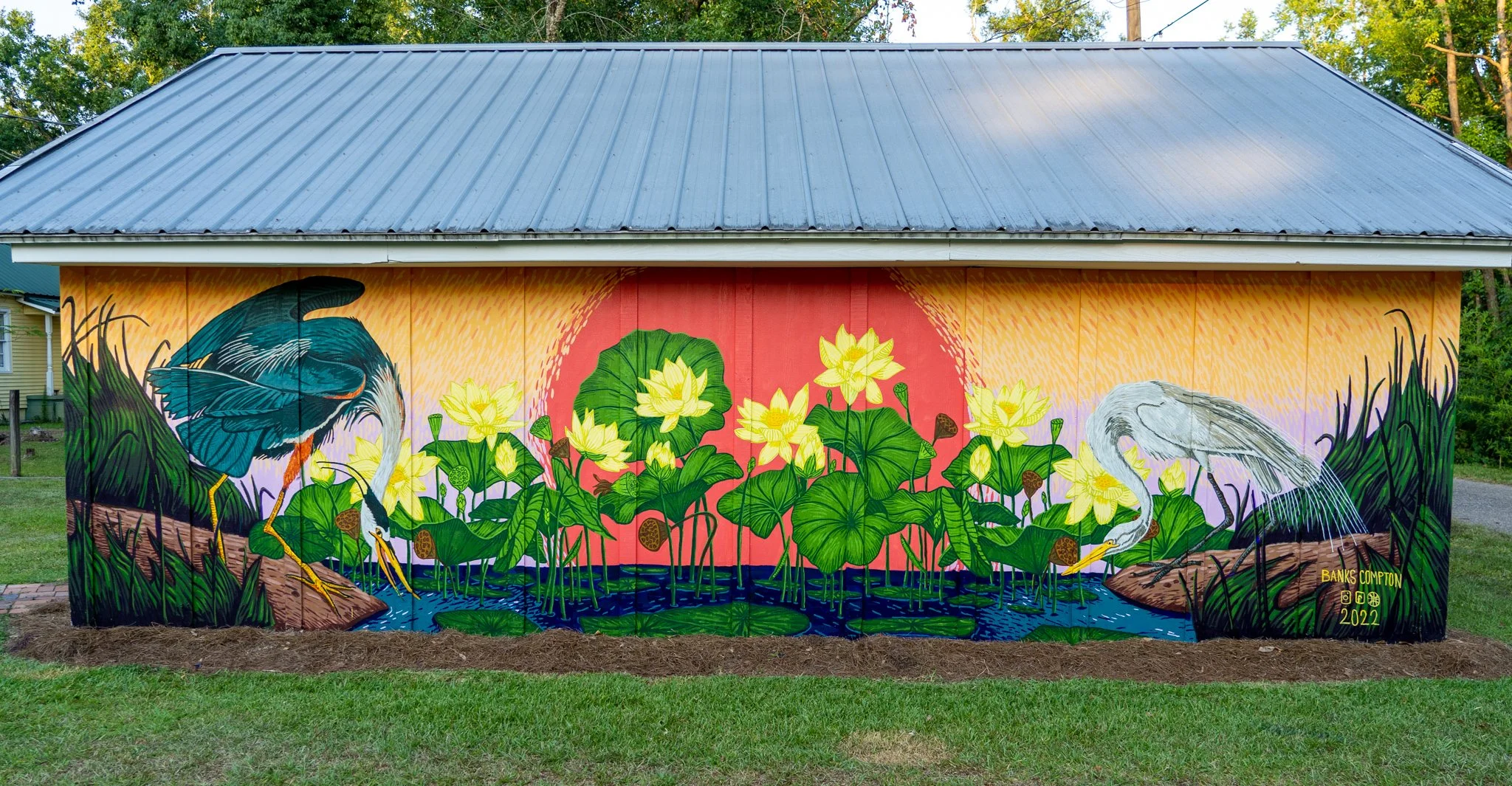 Exterior mural on Banks Compton's art studio in Foley, Alabama, featuring great blue herons, American lotus flowers, wetlands, and a colorful sunset.