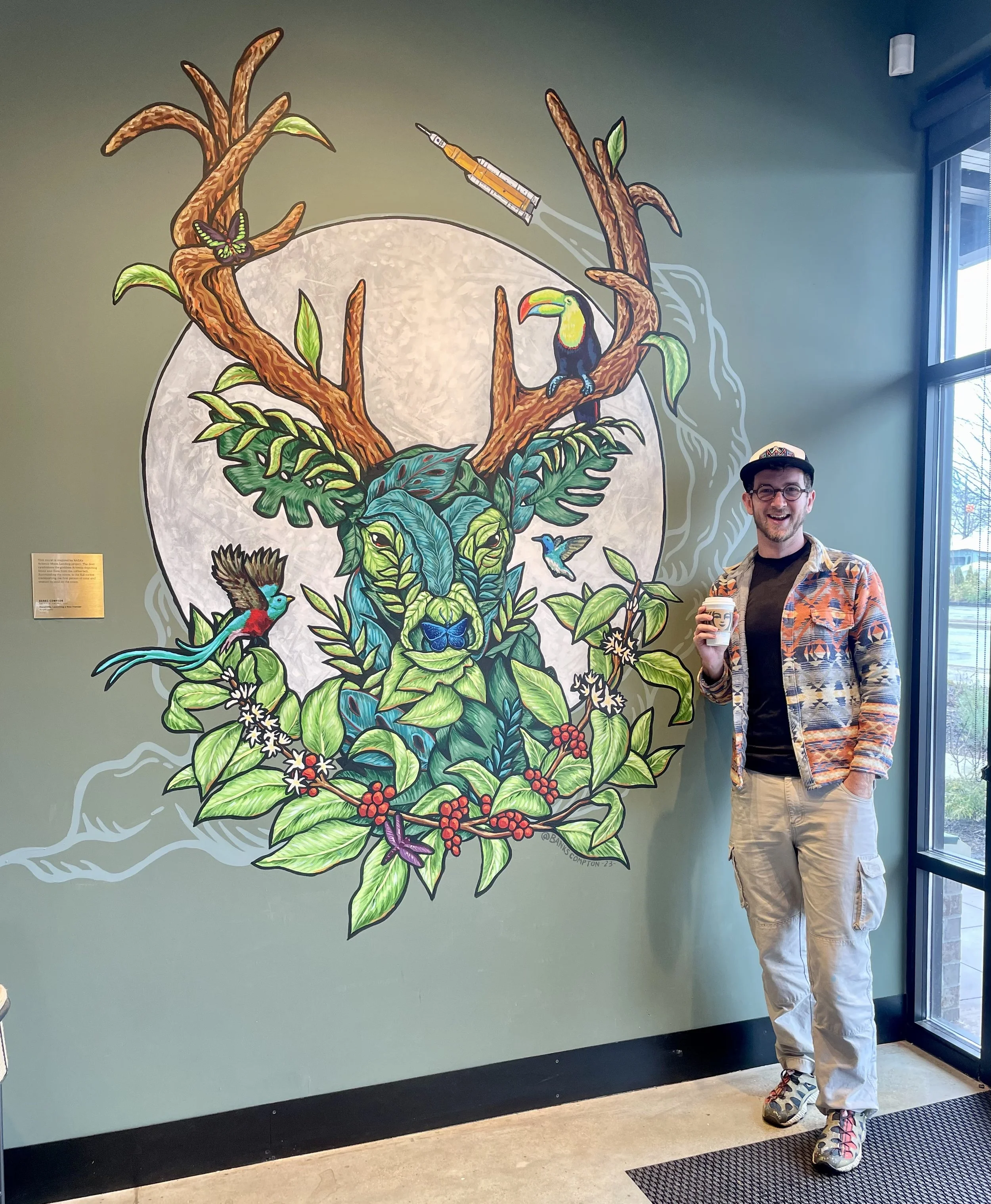 A man wearing glasses, a colorful patterned jacket, and beige cargo pants stands smiling in front of a vibrant mural of a tropical scene, holding a coffee cup. The mural features green foliage, a toucan, hummingbirds, butterflies, a tree with a syringe, and a large moon in the background.